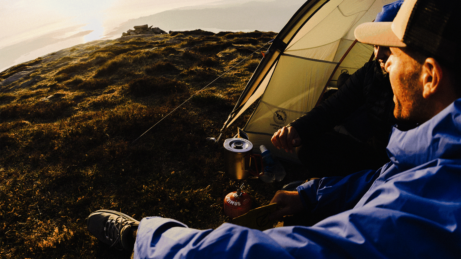 Two campers in jackets enjoy sunrise by a tent on a grassy hill, with a portable stove in the foreground.