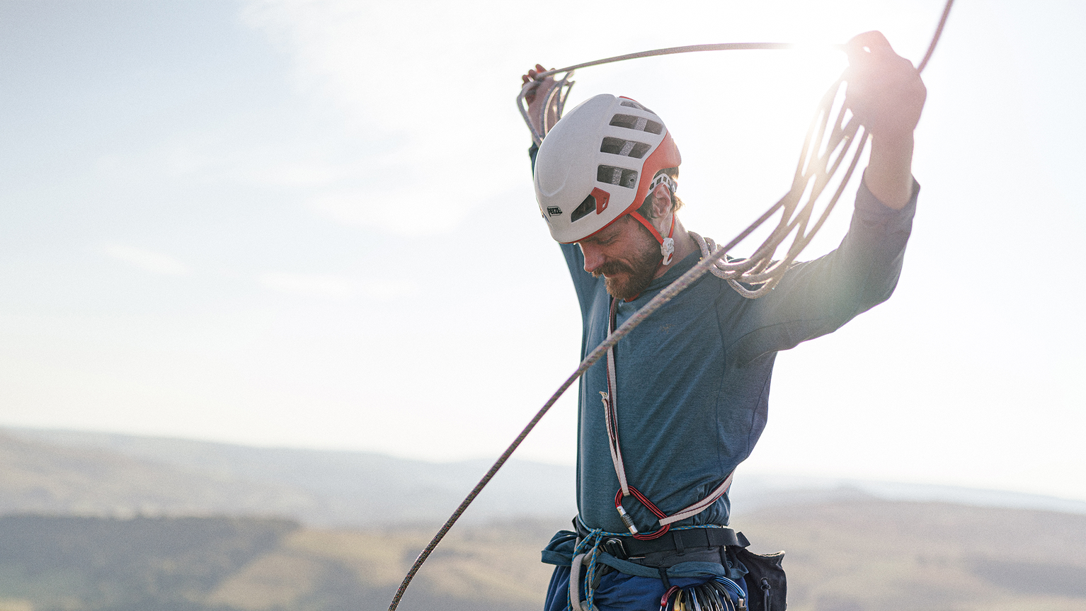 A climber wearing a helmet and harness organizes a rope against a bright sky, standing on a rocky landscape.