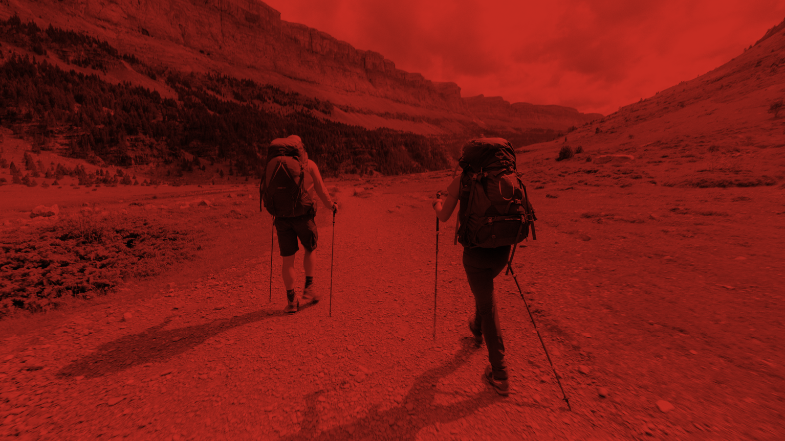 Two hikers with backpacks and trekking poles walking along a rugged mountain trail.