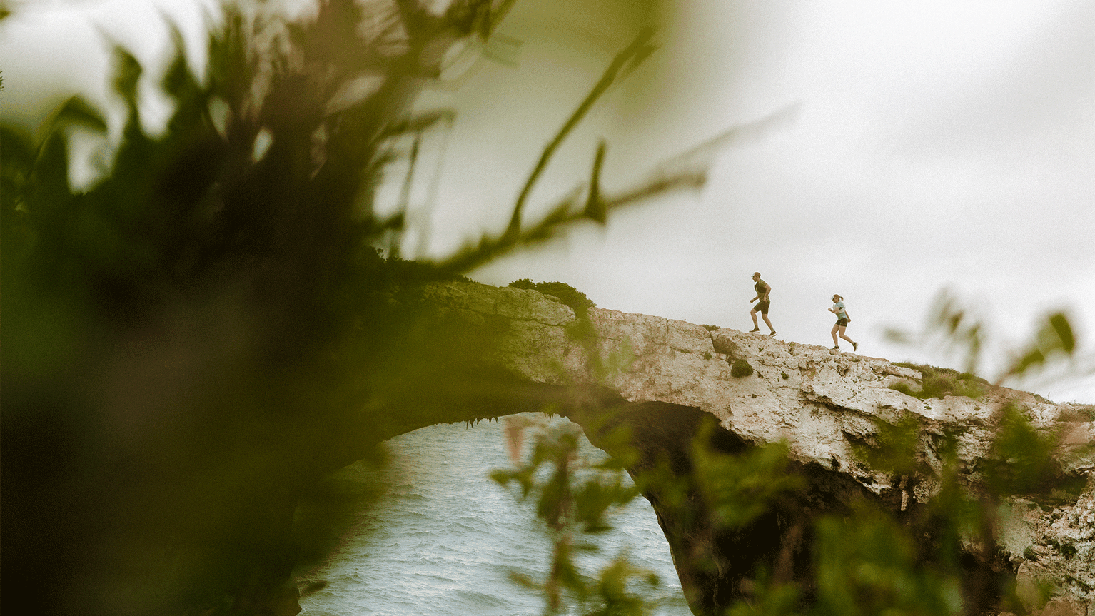 Two people running on a rocky arch over the sea, framed by blurred green foliage.