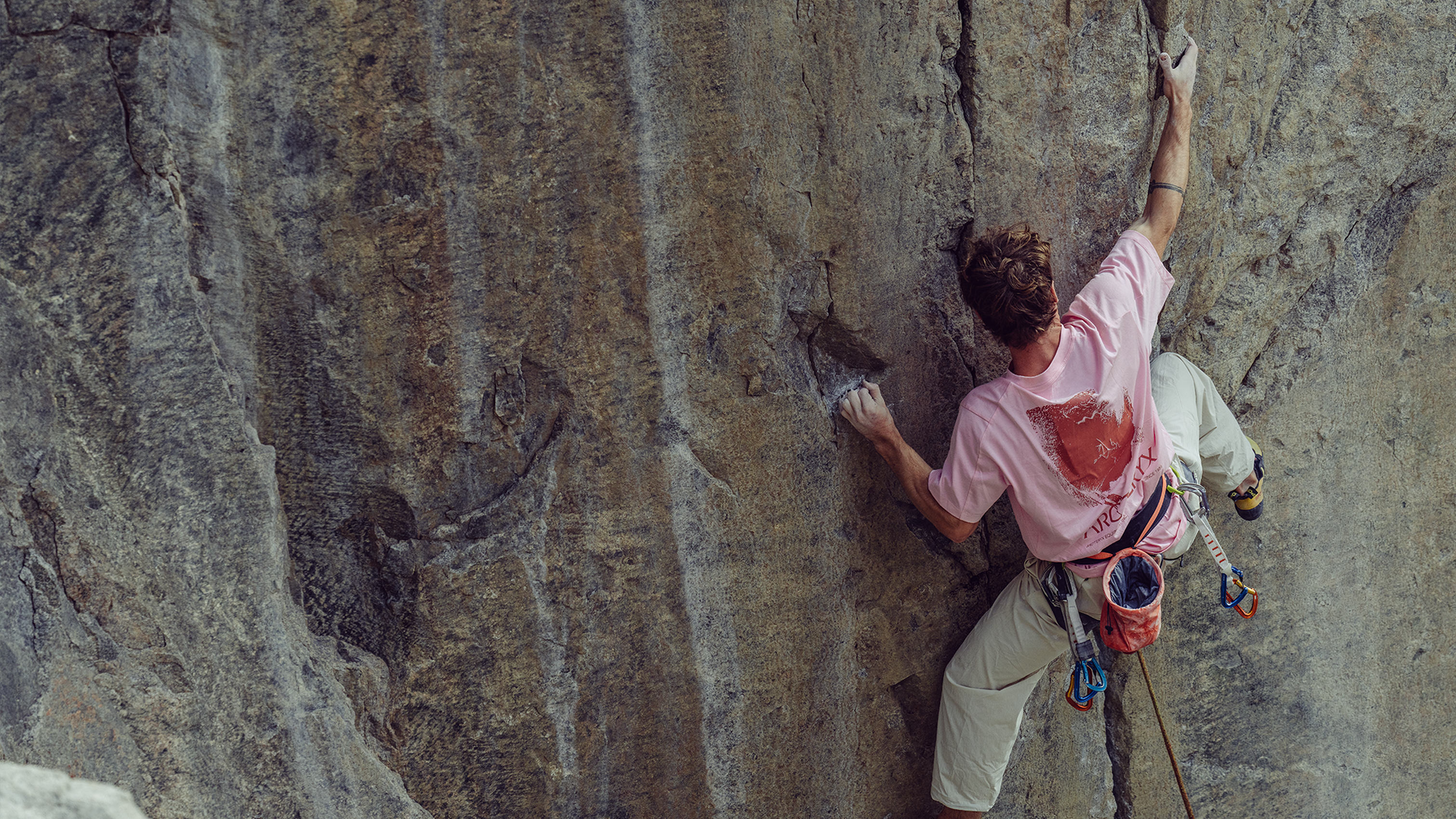 Climber tackles a challenging rock face, using safety gear. Wears a pink shirt and white trousers, showcasing mountain sports expertise.