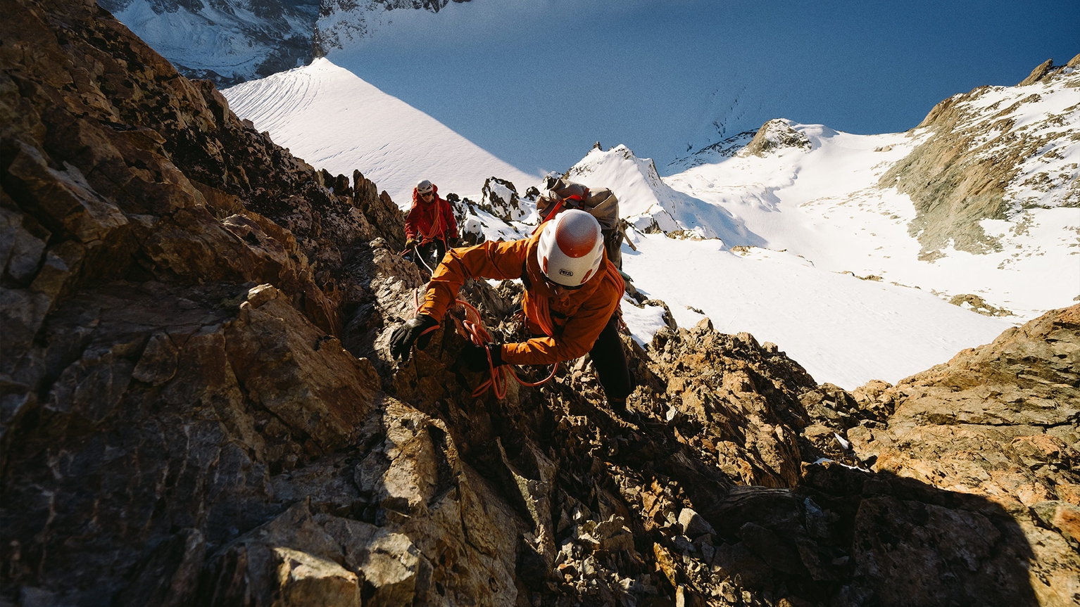 Climbers wearing helmets and gear ascend a steep, rocky mountain with snowy peaks in the background under a clear blue sky.