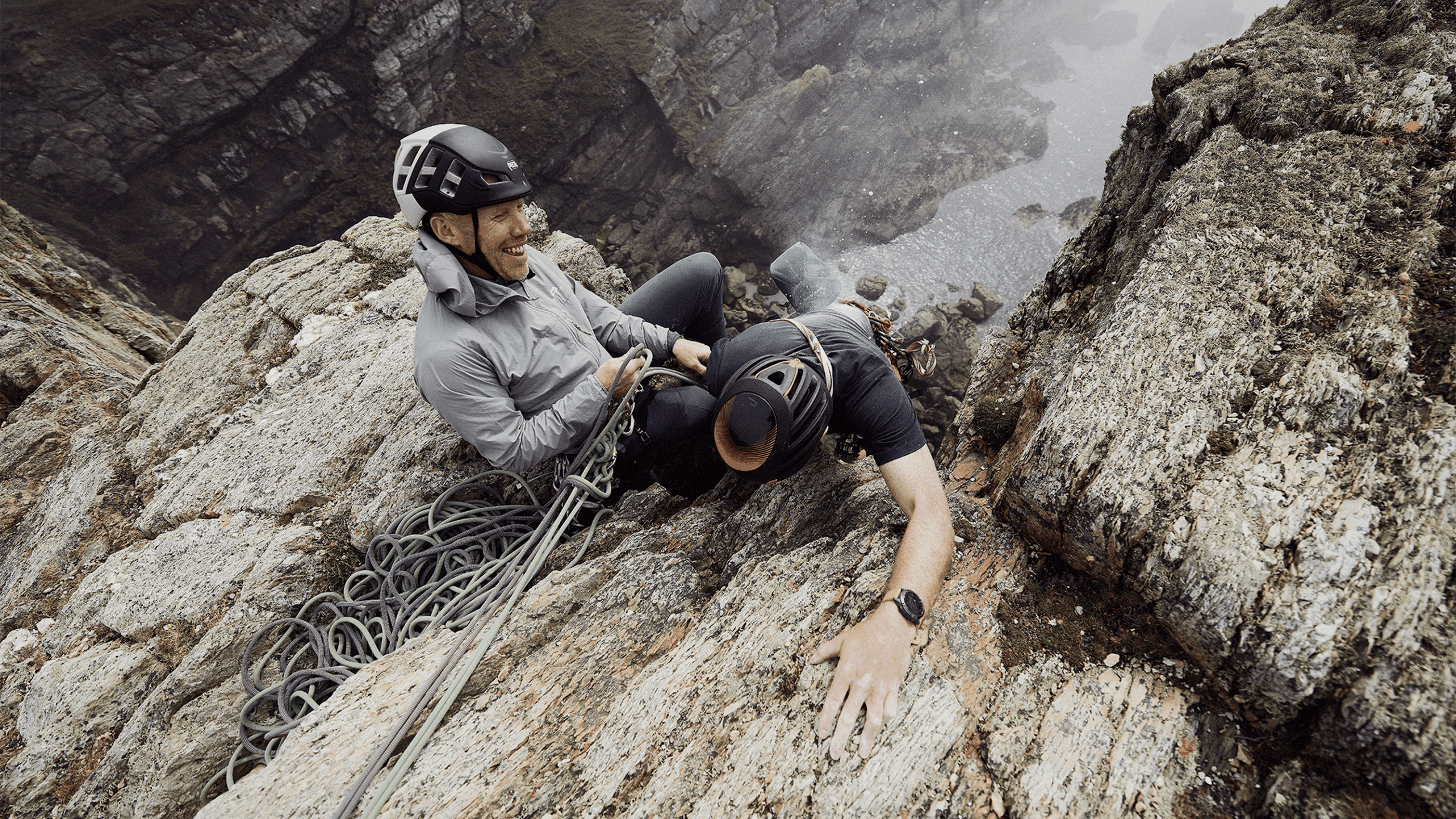 Two climbers on a rugged cliffside, with one assisting the other. Steep drop and ocean visible below. Both are wearing helmets and gear.
