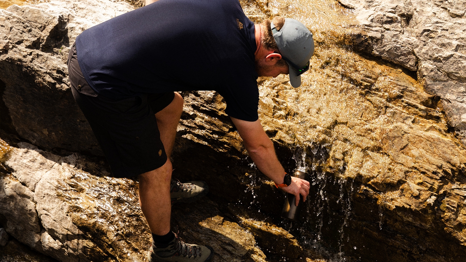 Man in outdoor gear fills water bottle from a mountain stream on rocky terrain.