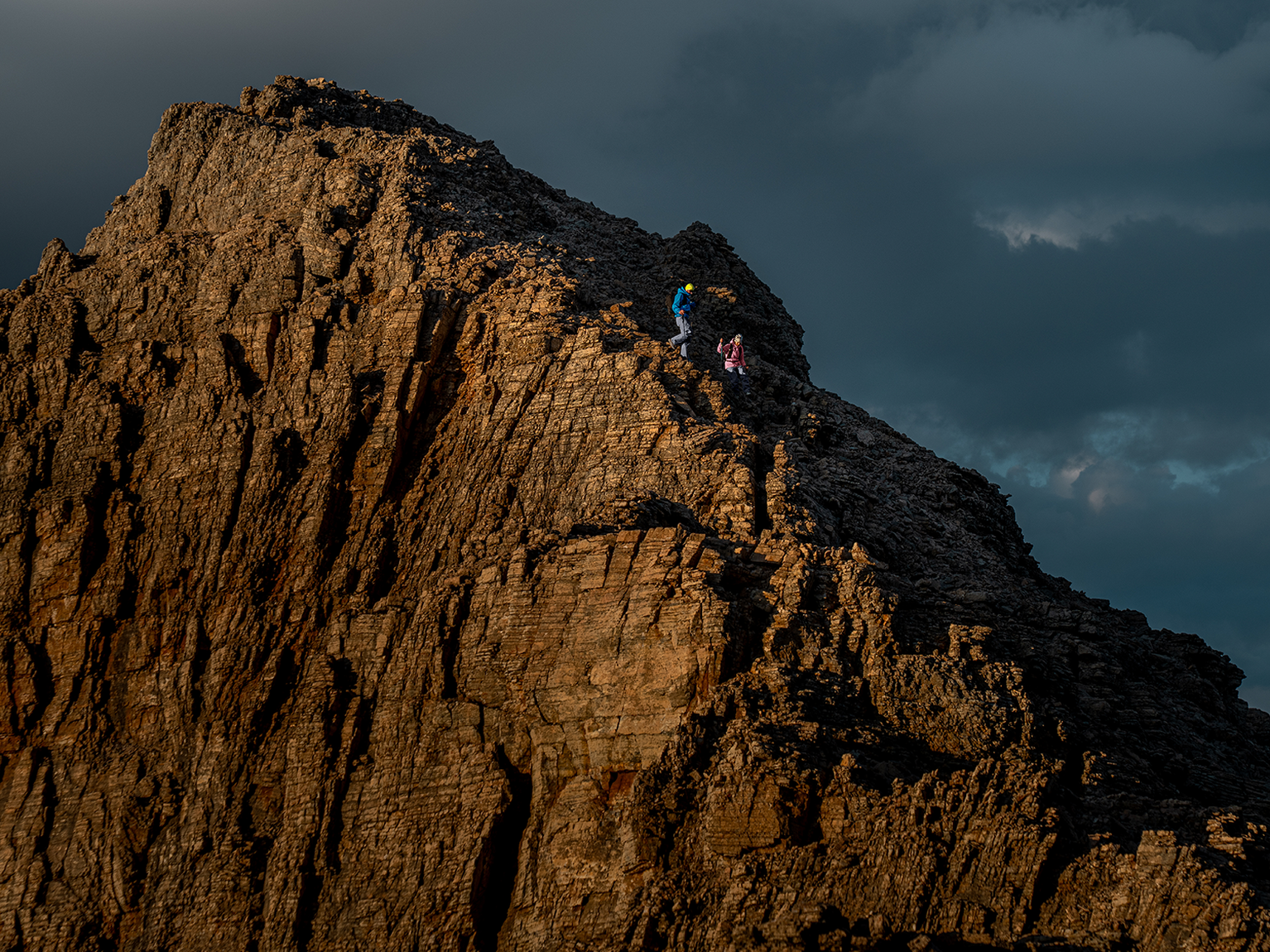 Two climbers traverse a rugged mountain ridge under a moody sky, highlighting the dramatic landscape and their daring journey.