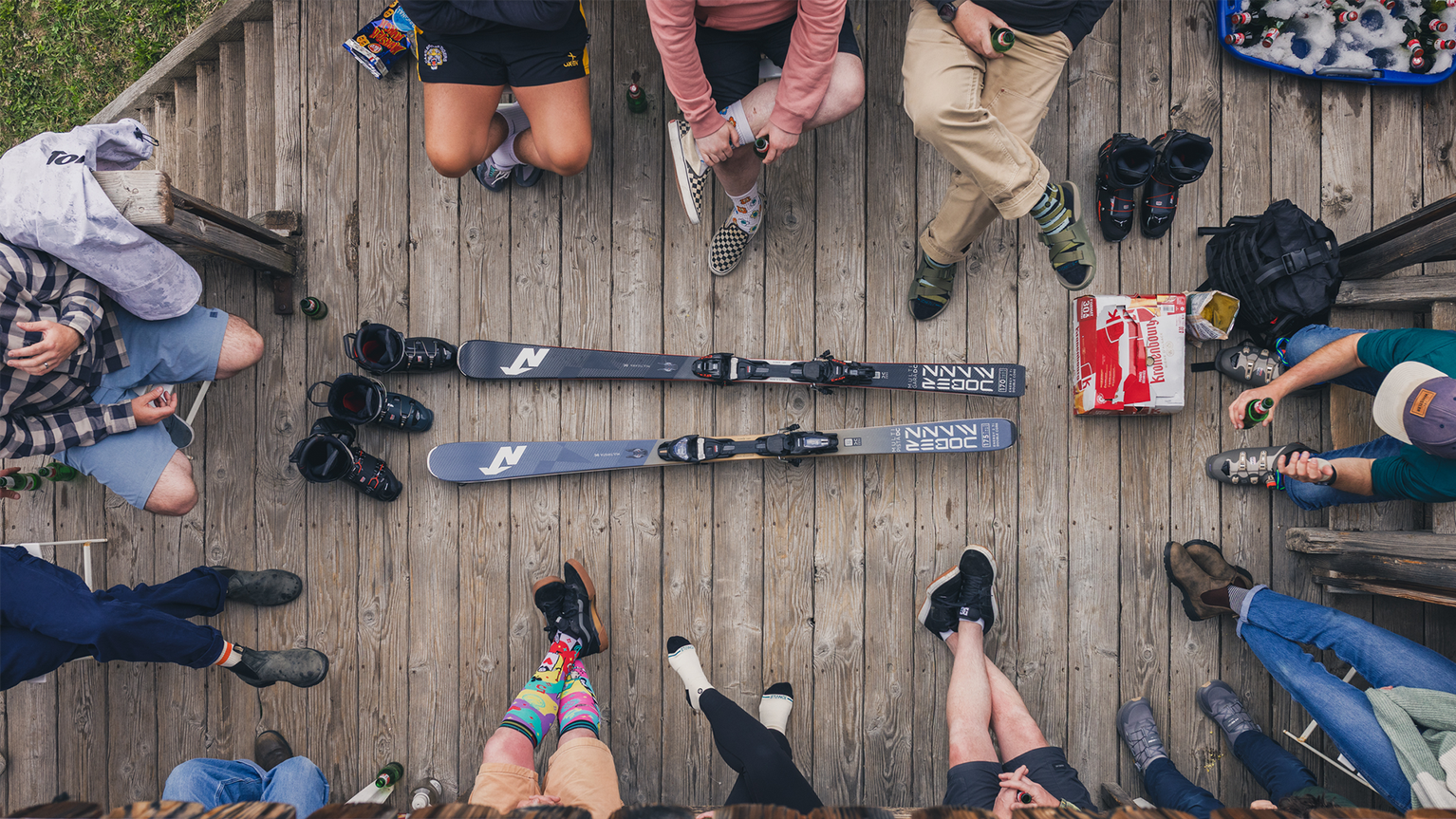 A group of people sitting on a wooden deck in a circle, with two skis in the center. Various shoes and colorful socks are visible.