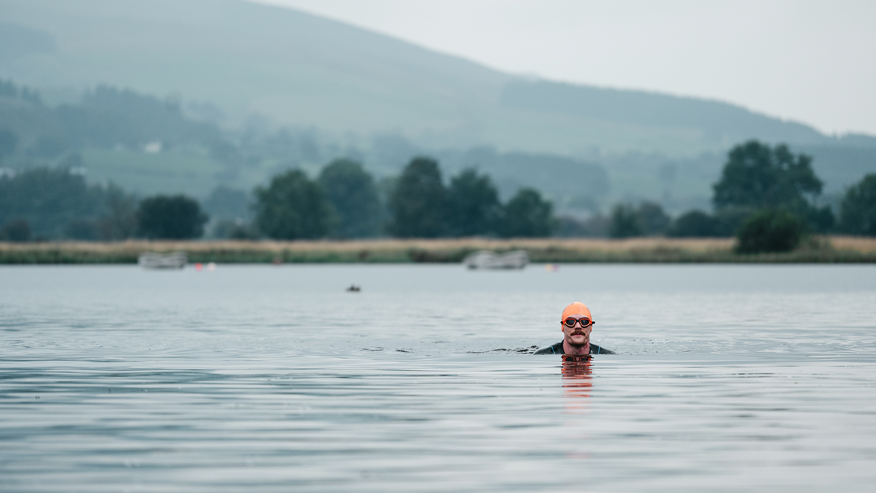 Person swimming in a lake, wearing orange goggles and cap, with blurred greenery in the background and a calm water surface.