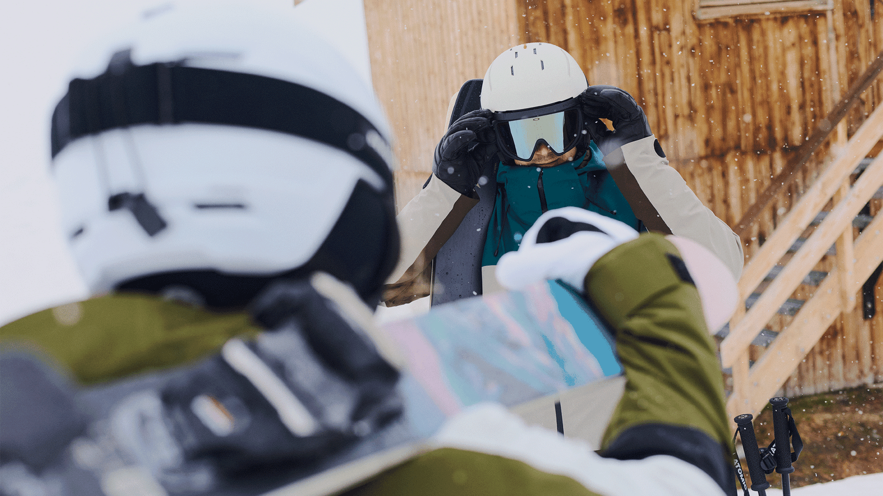 Two skiers in winter gear, one adjusting goggles while holding skis.