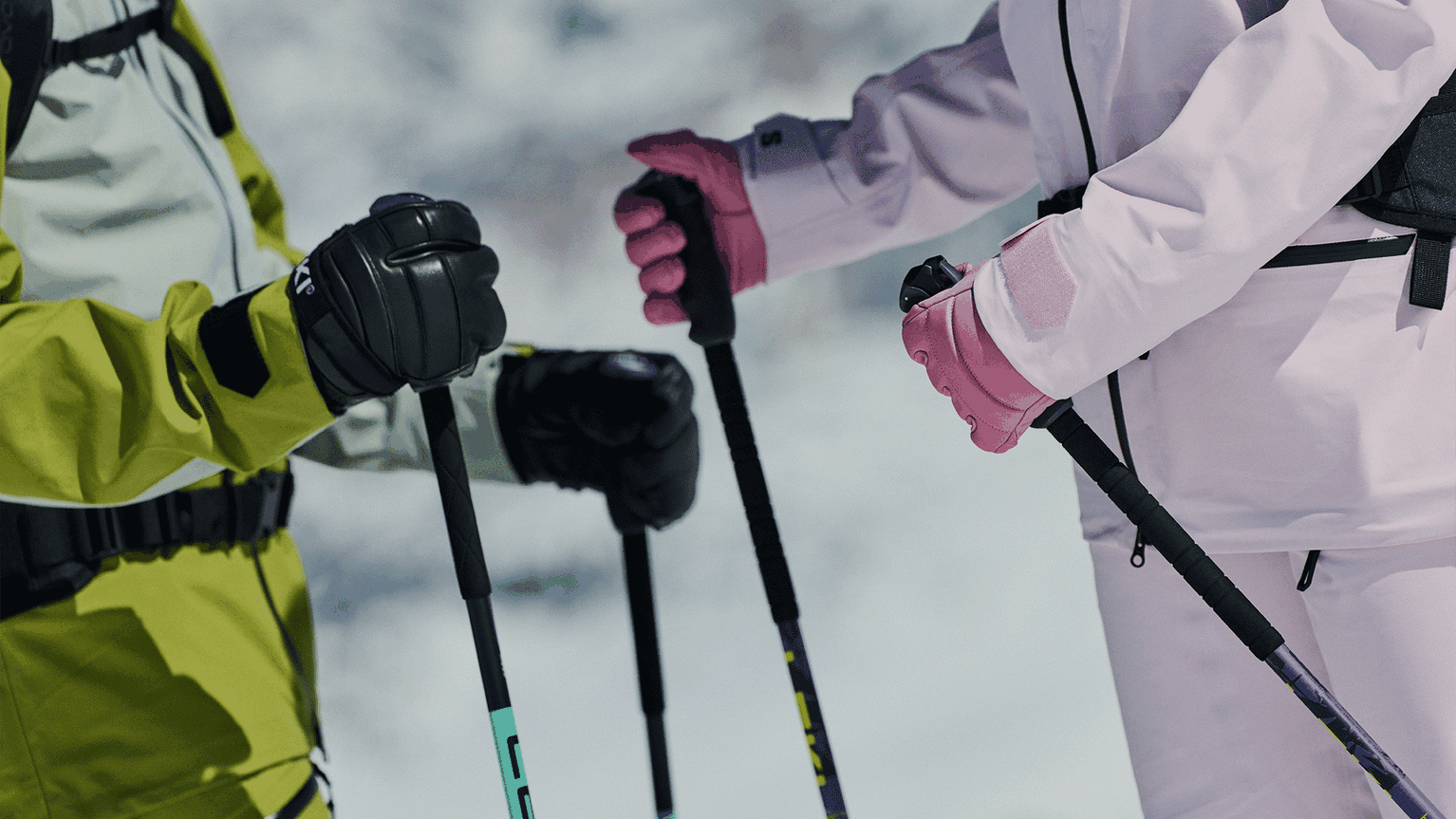 Two people wearing ski gear and gloves, holding colourful ski poles against a snowy background.