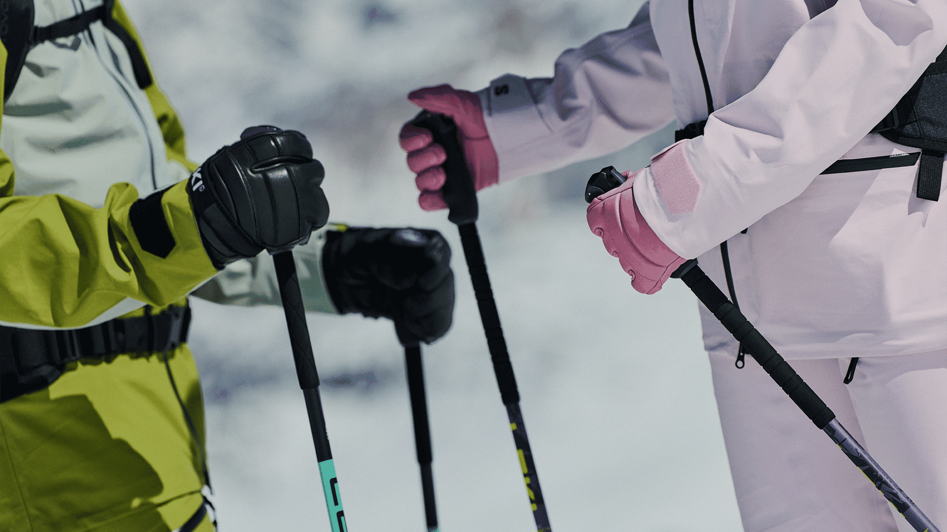 Two people wearing ski gear and gloves, holding colourful ski poles against a snowy background.
