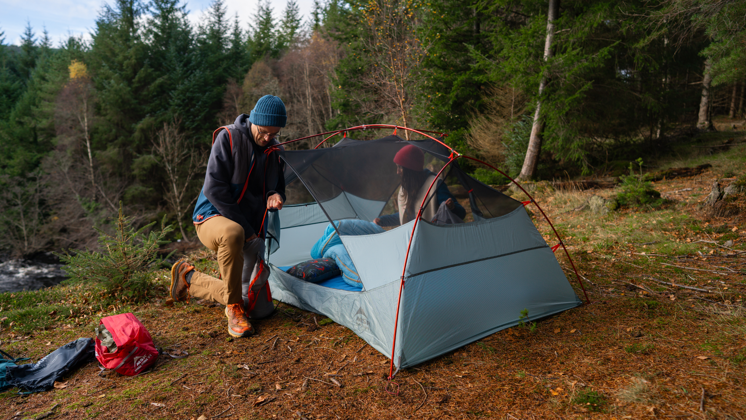 Man setting up a tent in a forest clearing, with two people inside the tent, surrounded by trees and a nearby stream.
