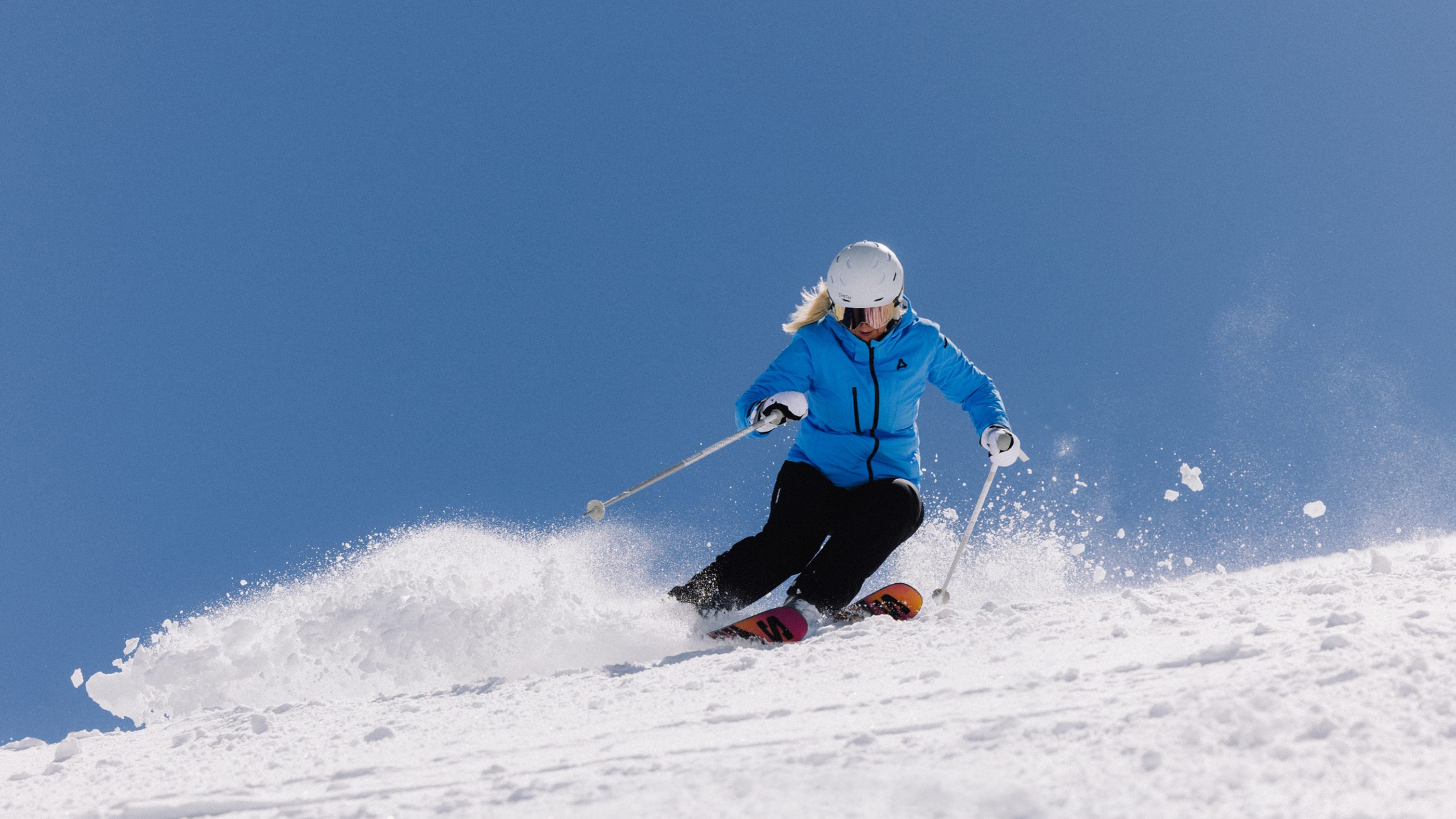 Skier in blue jacket and white helmet carving through powder on sunny slope, high-performance skis and poles.