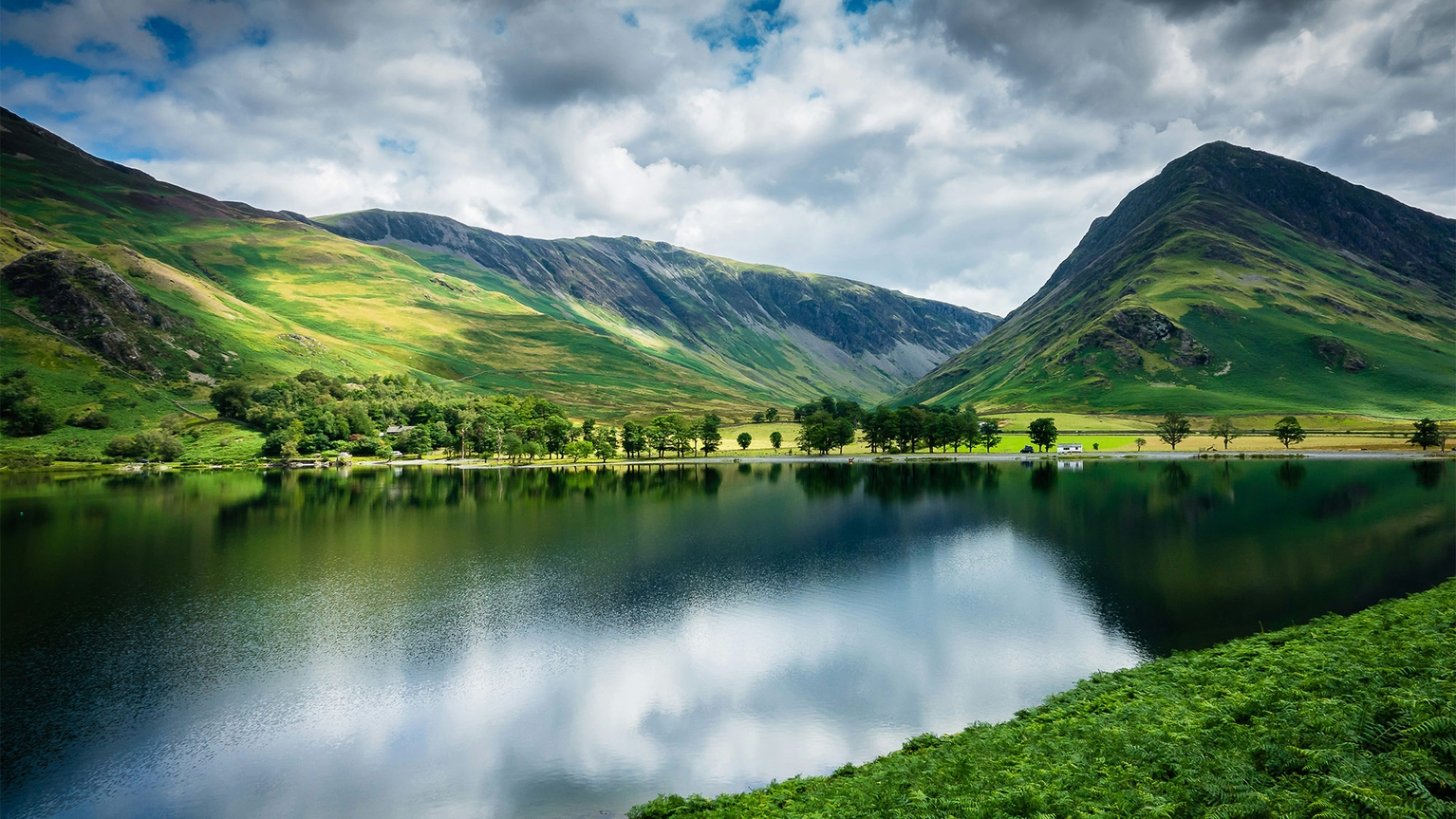 A serene lake reflects lush green hills and a cloudy sky, with a line of trees along the shore and mountains in the background.