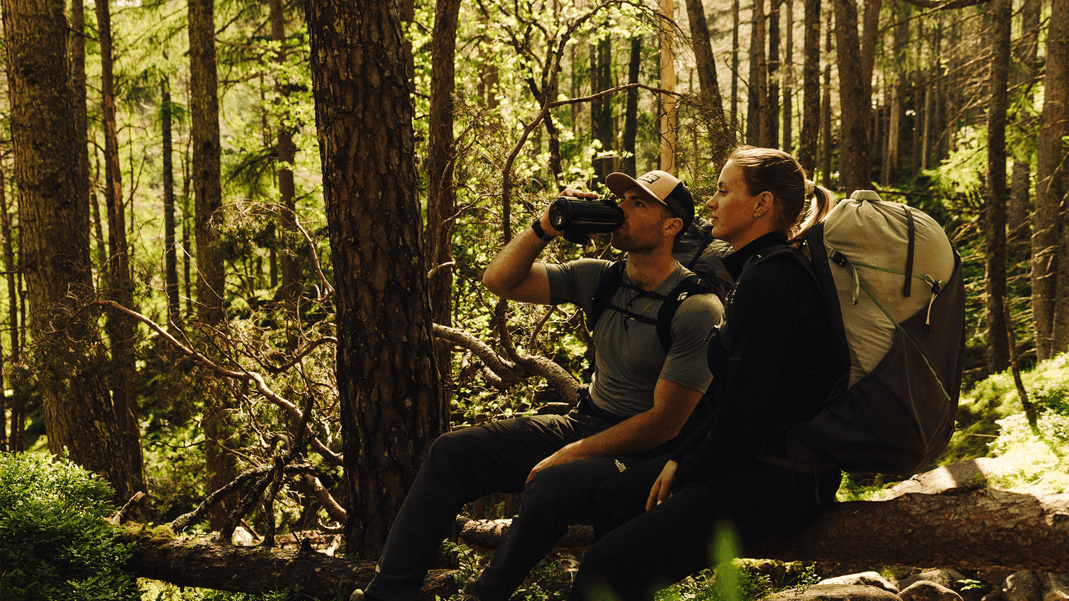 Two hikers rest on a log in a sunlit forest, one drinking from a bottle. Both wear backpacks and outdoor clothing, enjoying a break.