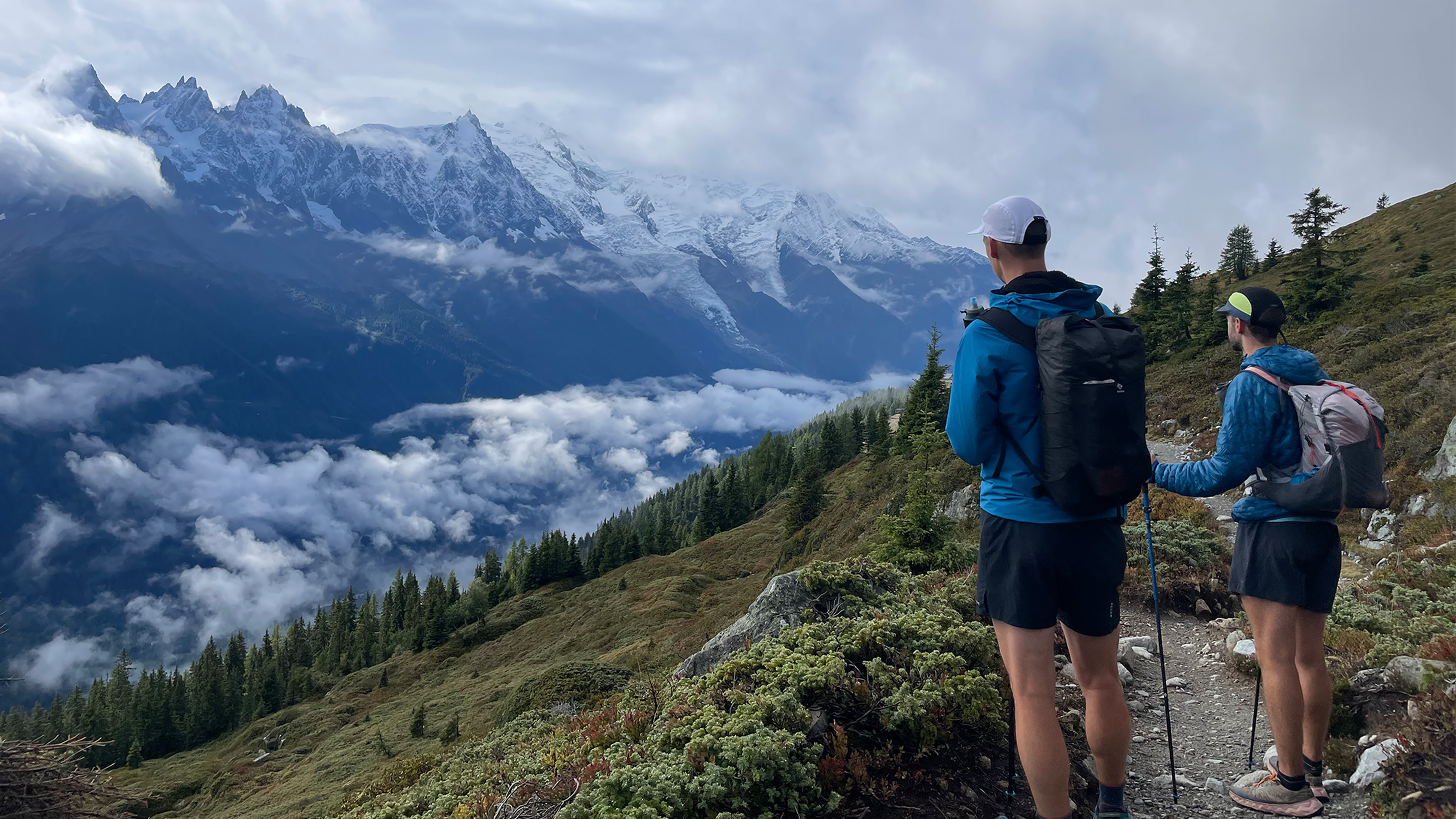 Two hikers with backpacks and trekking poles admire a scenic mountain view, surrounded by clouds and snow-capped peaks.