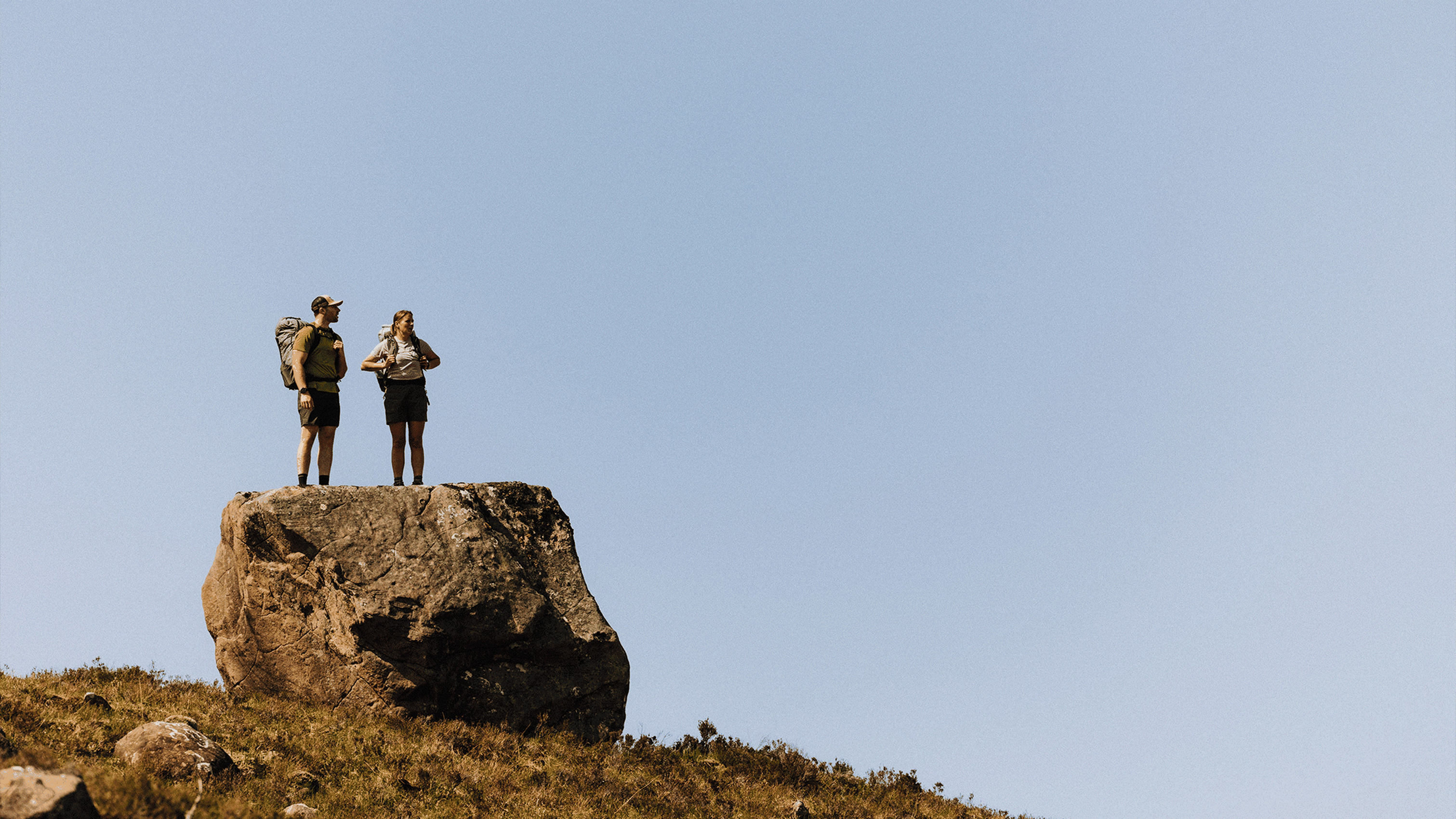 Two hikers with backpacks stand on a large rock, enjoying a clear, blue sky view.