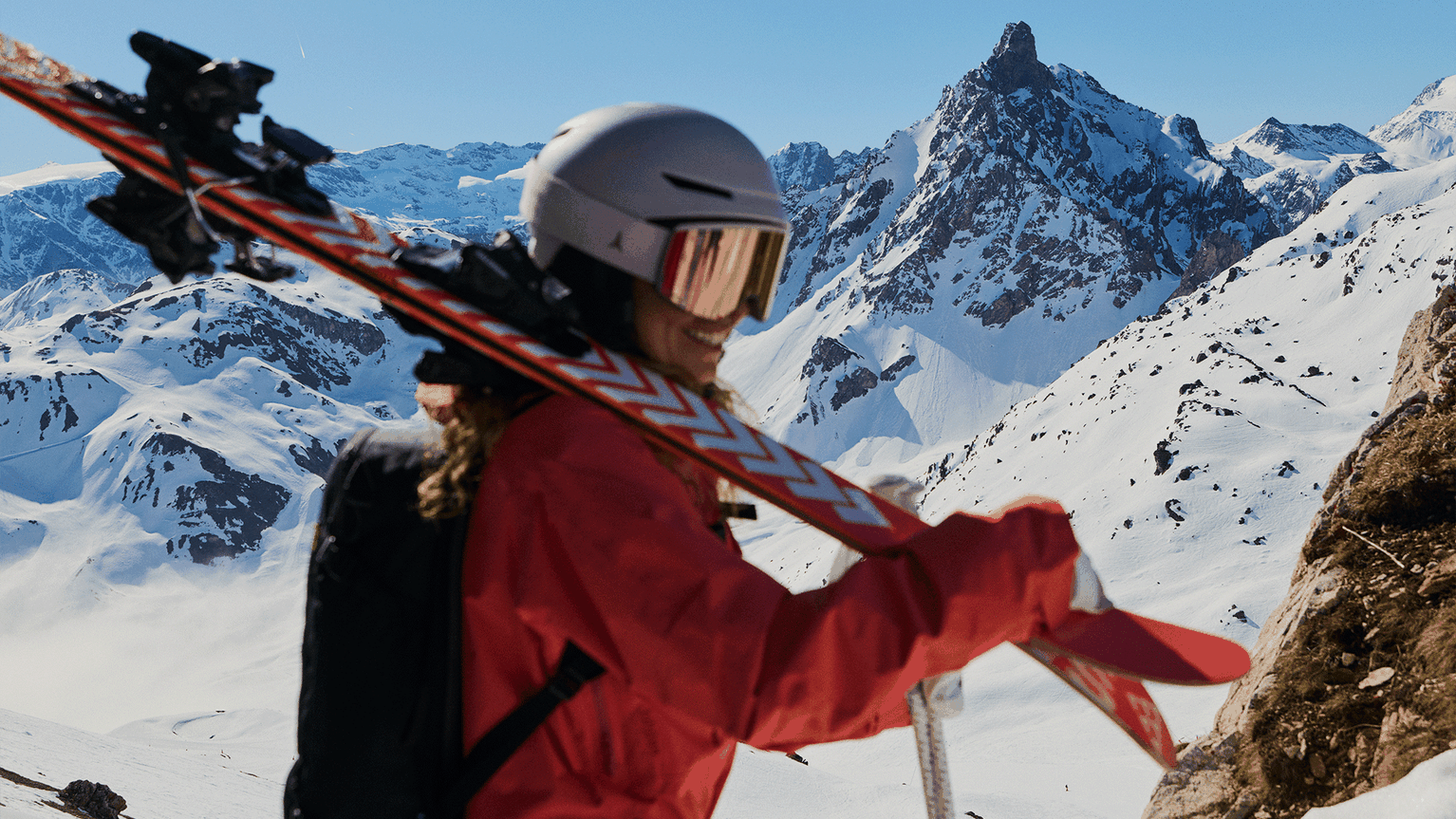Smiling skier in red jacket and helmet carries skis on snow-covered mountain landscape background.