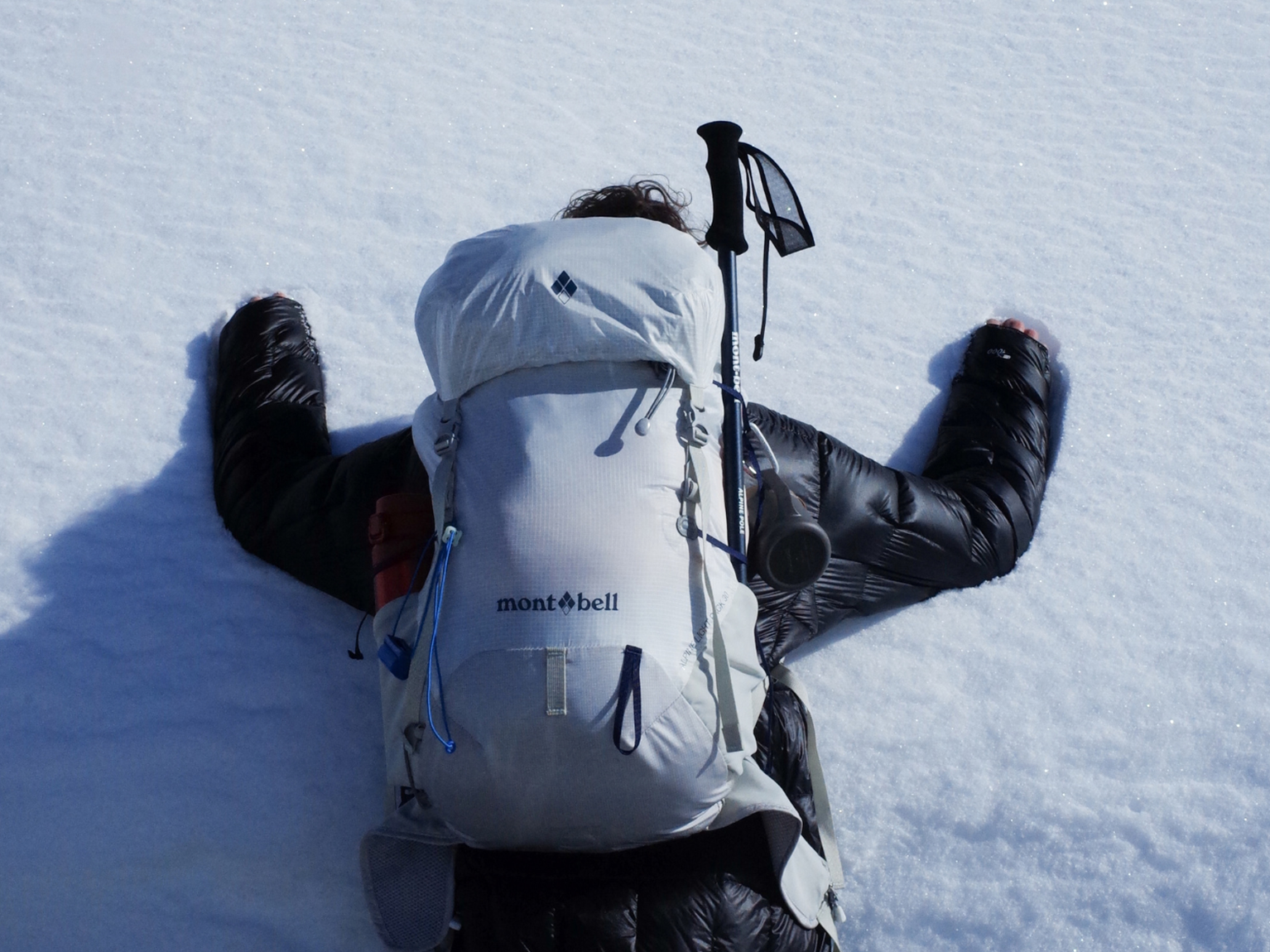 Person face down in the snow with a large backpack and trekking pole attached, wearing a black puffer jacket.