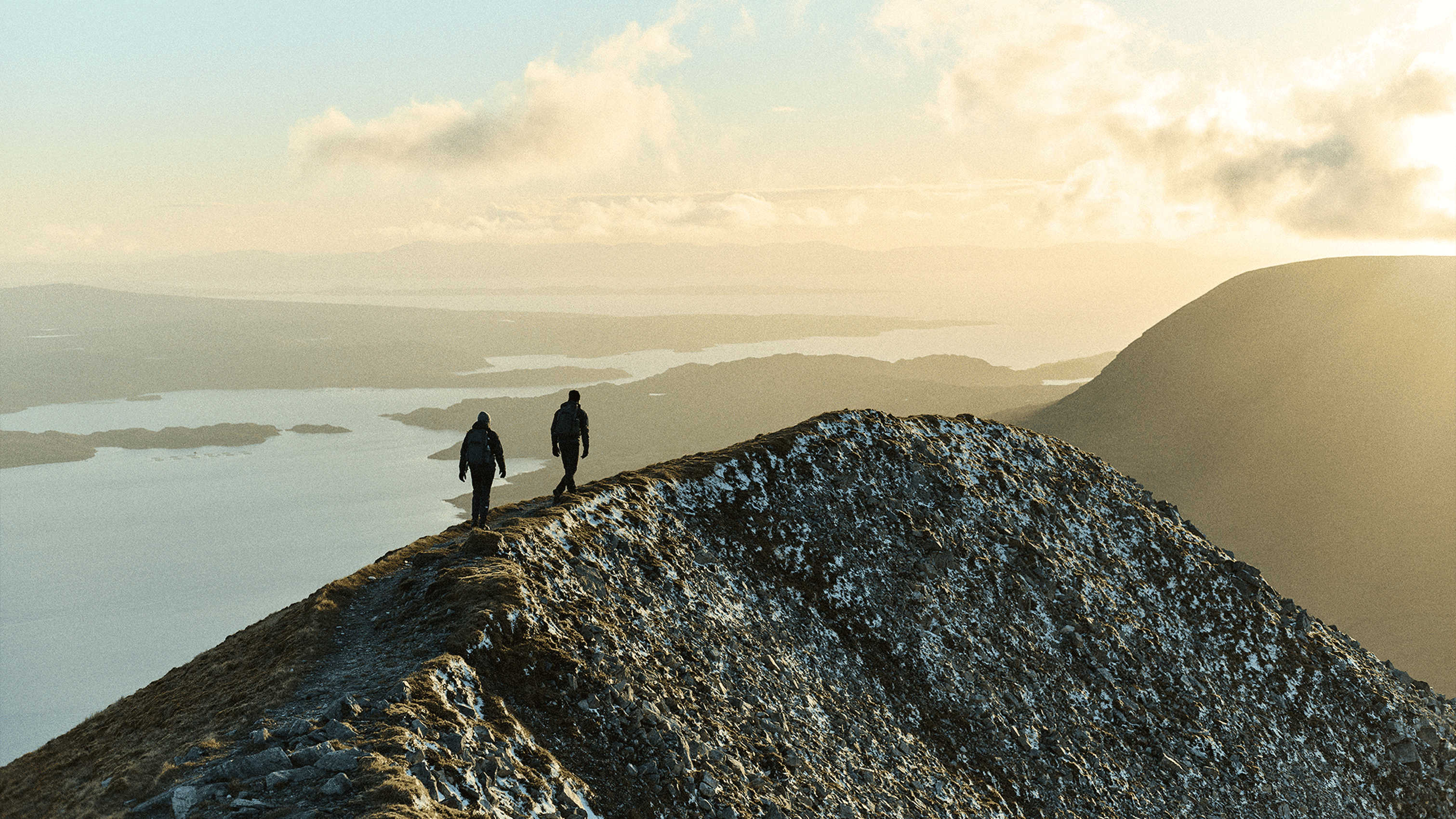 Two hikers walk along a snowy mountain ridge at sunrise, overlooking a vast landscape of lakes and distant hills.
