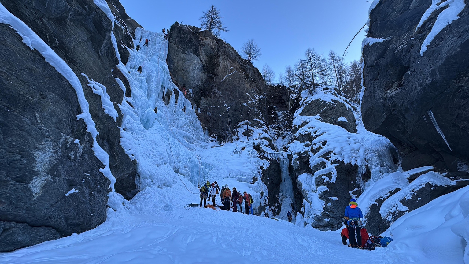 Group of people ice climbing on a frozen waterfall amid snowy cliffs, with some climbers preparing at the base and others on the climb.