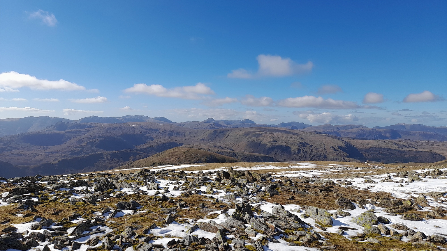 Snow-dusted rocky landscape with distant rolling hills under a bright blue sky and scattered clouds.