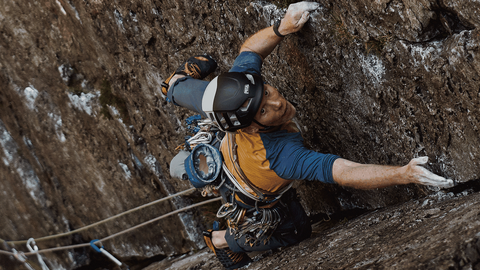 Climber in a helmet scaling a steep rock face, gripping tightly with chalked hands, wearing a harness equipped with climbing gear.