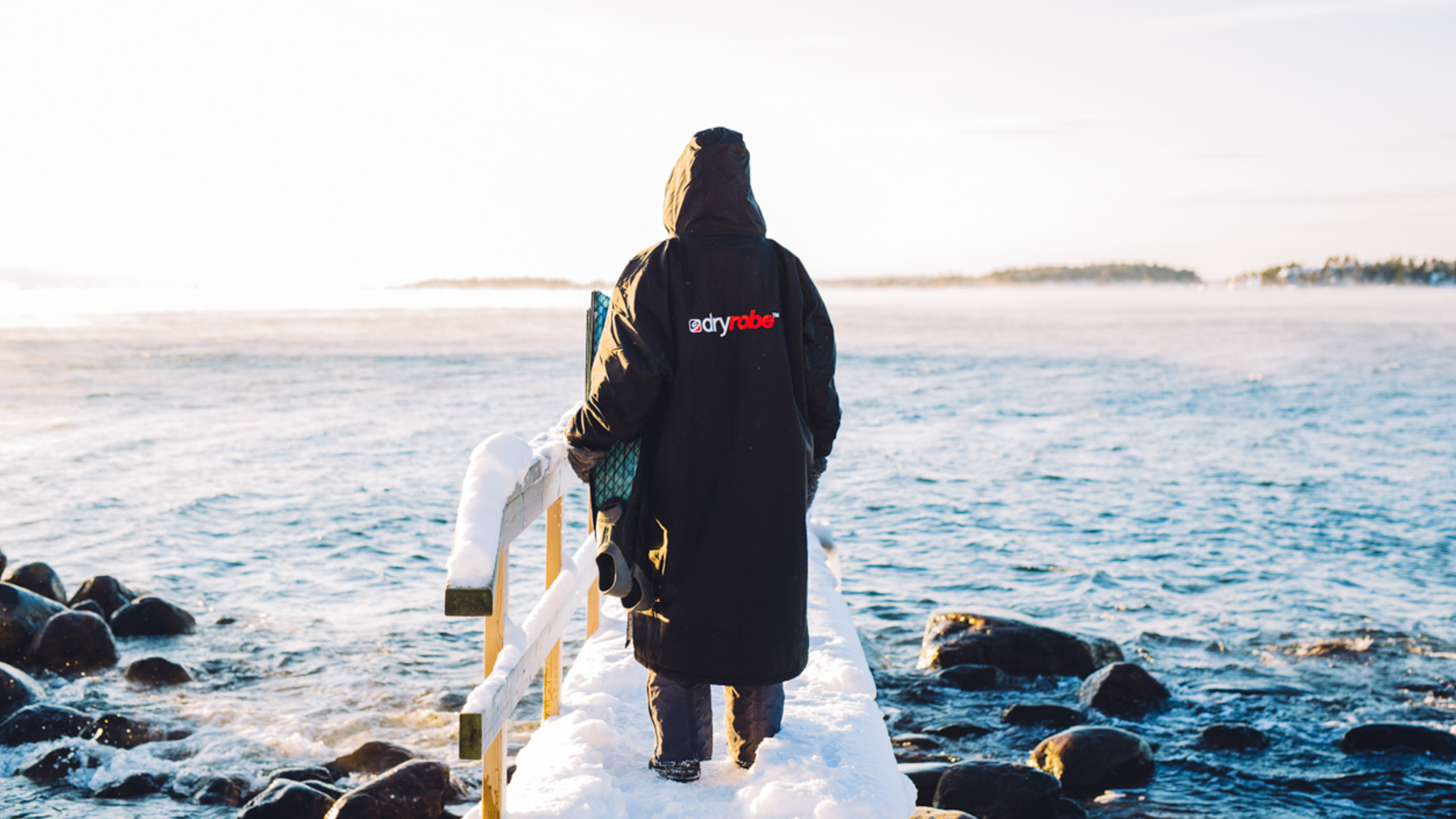 Person in a dryrobe stands on a snowy pier, overlooking a calm, expansive sea under a clear sky.