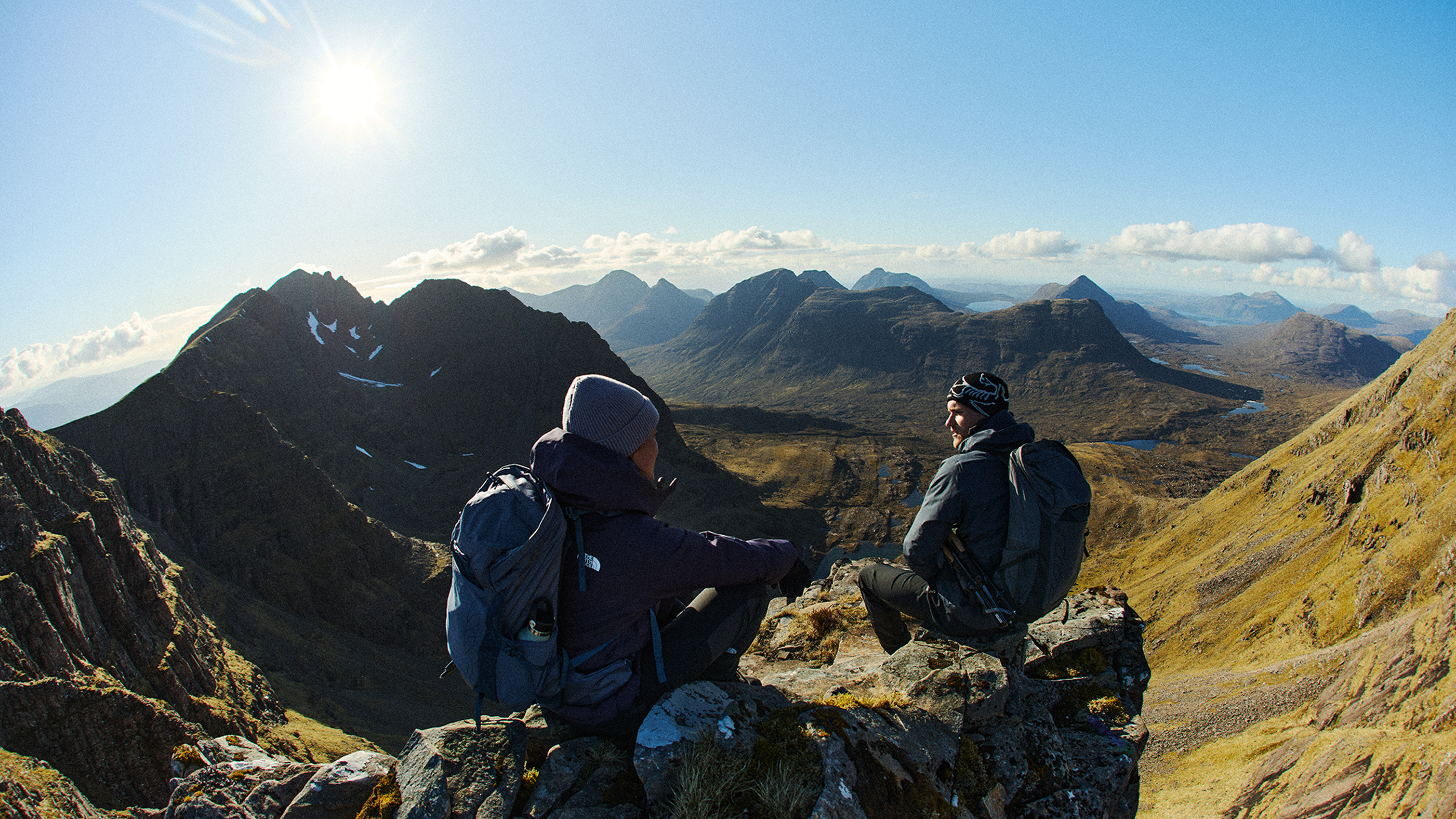 Two hikers in warm gear rest on a rocky peak, overlooking a vast mountain range under a clear, bright sky.