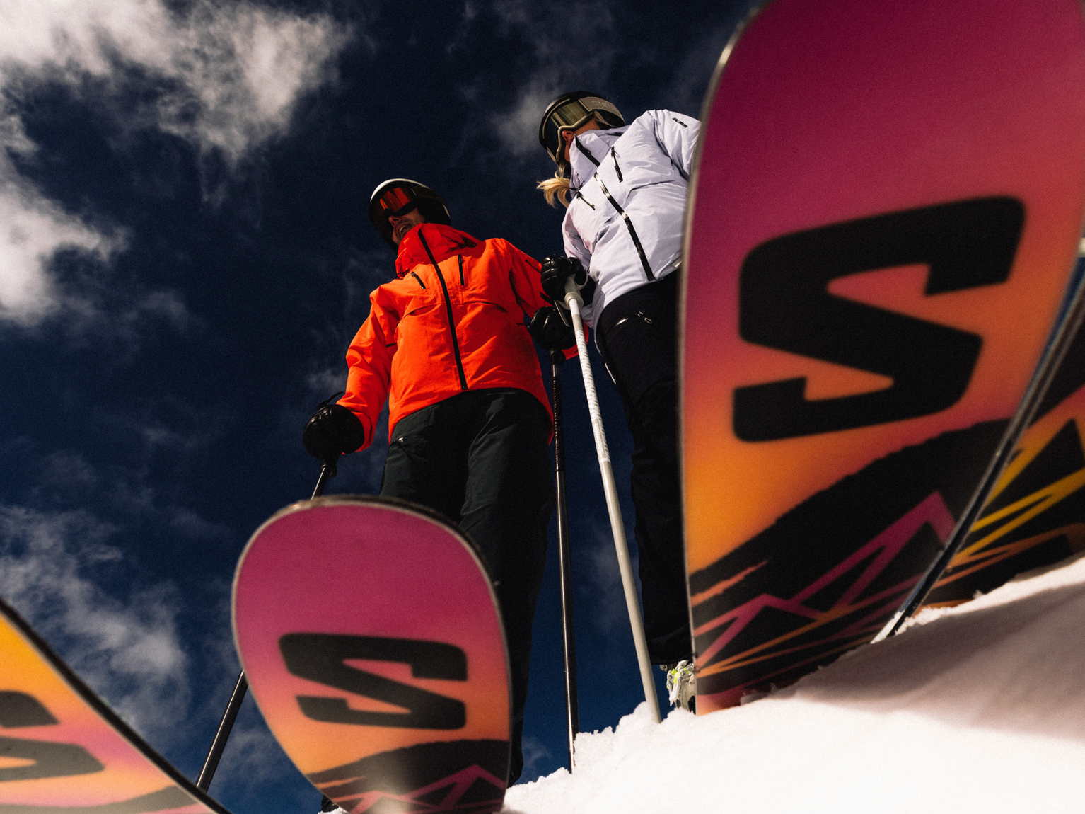 Two skiers in bright jackets stand on a snowy slope, holding poles, with colourful skis in the foreground against a clear blue sky.