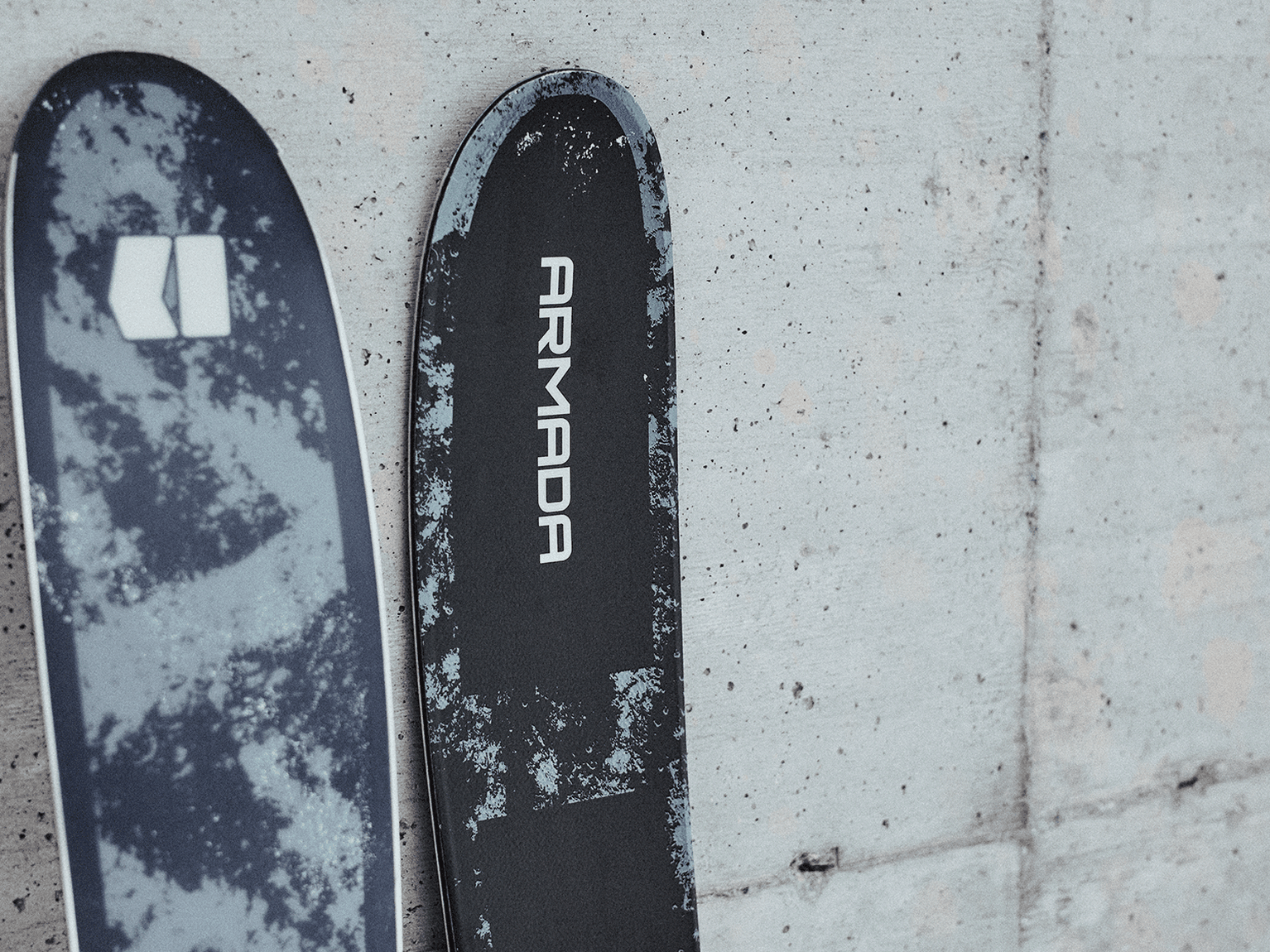Close-up of two ski tips, featuring contrasting patterns and the brand name "Armada" on one of them, against a textured concrete background.
