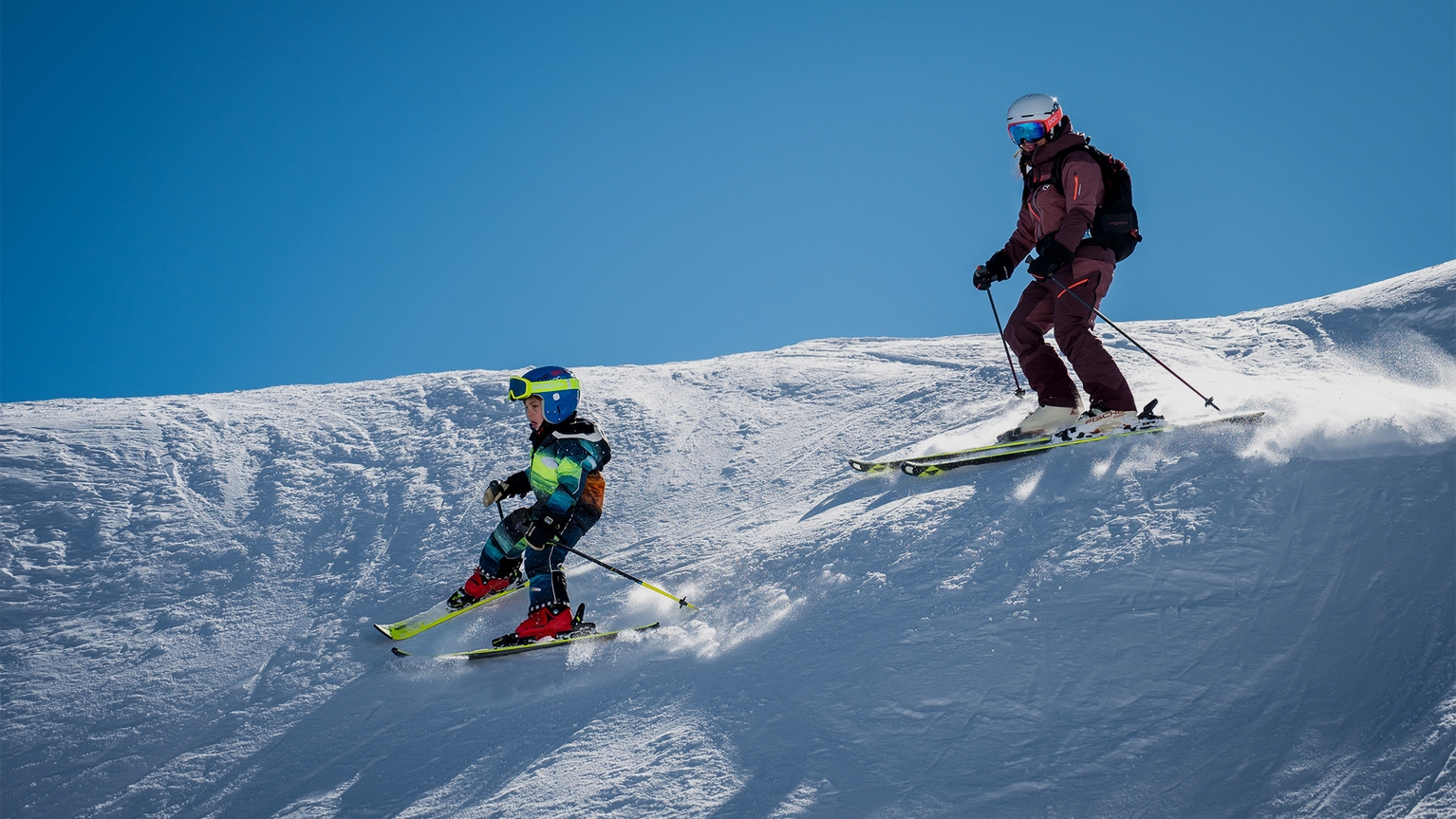 Adult and child skiing downhill on a snowy mountain slope under a clear blue sky, showcasing dynamic movement and vibrant gear.
