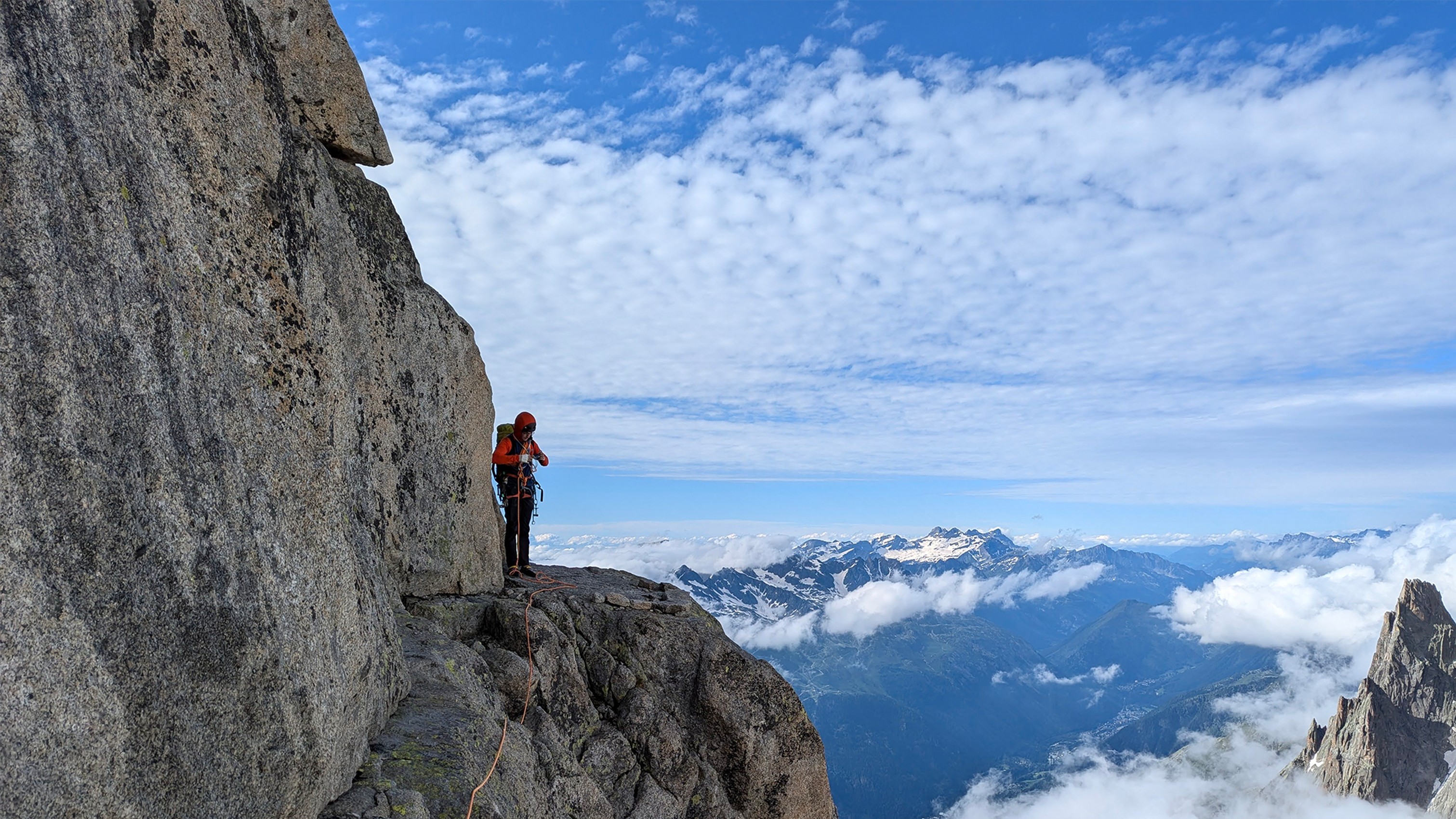A climber in an orange jacket stands on a rocky ledge with a vast mountain range and cloudy sky in the background.