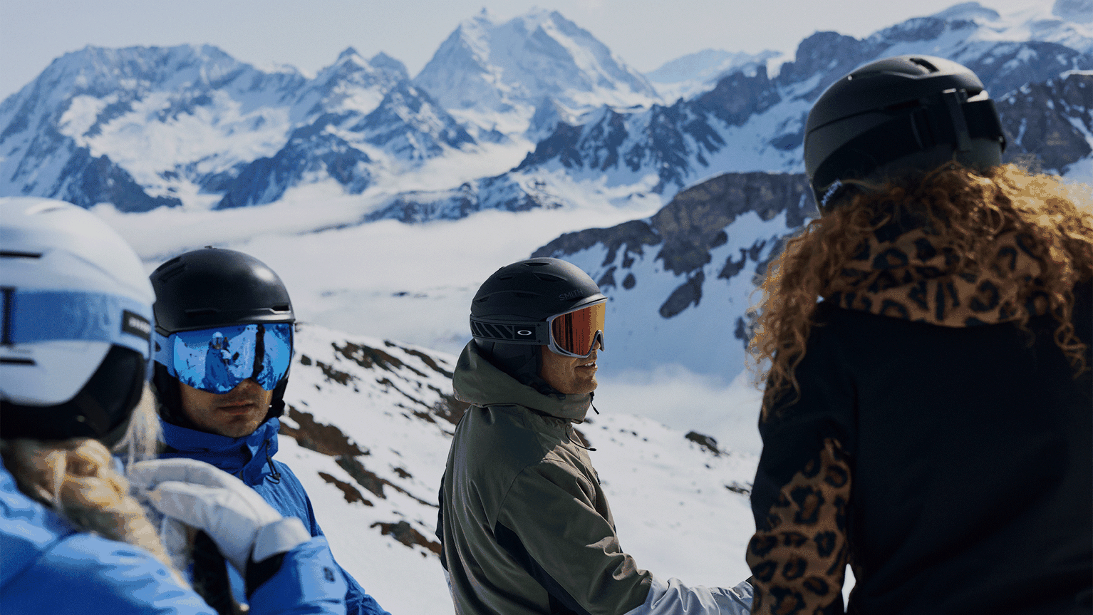 Skiers chatting on a snowy mountain slope, wearing helmets and goggles, with stunning peaks in the background.