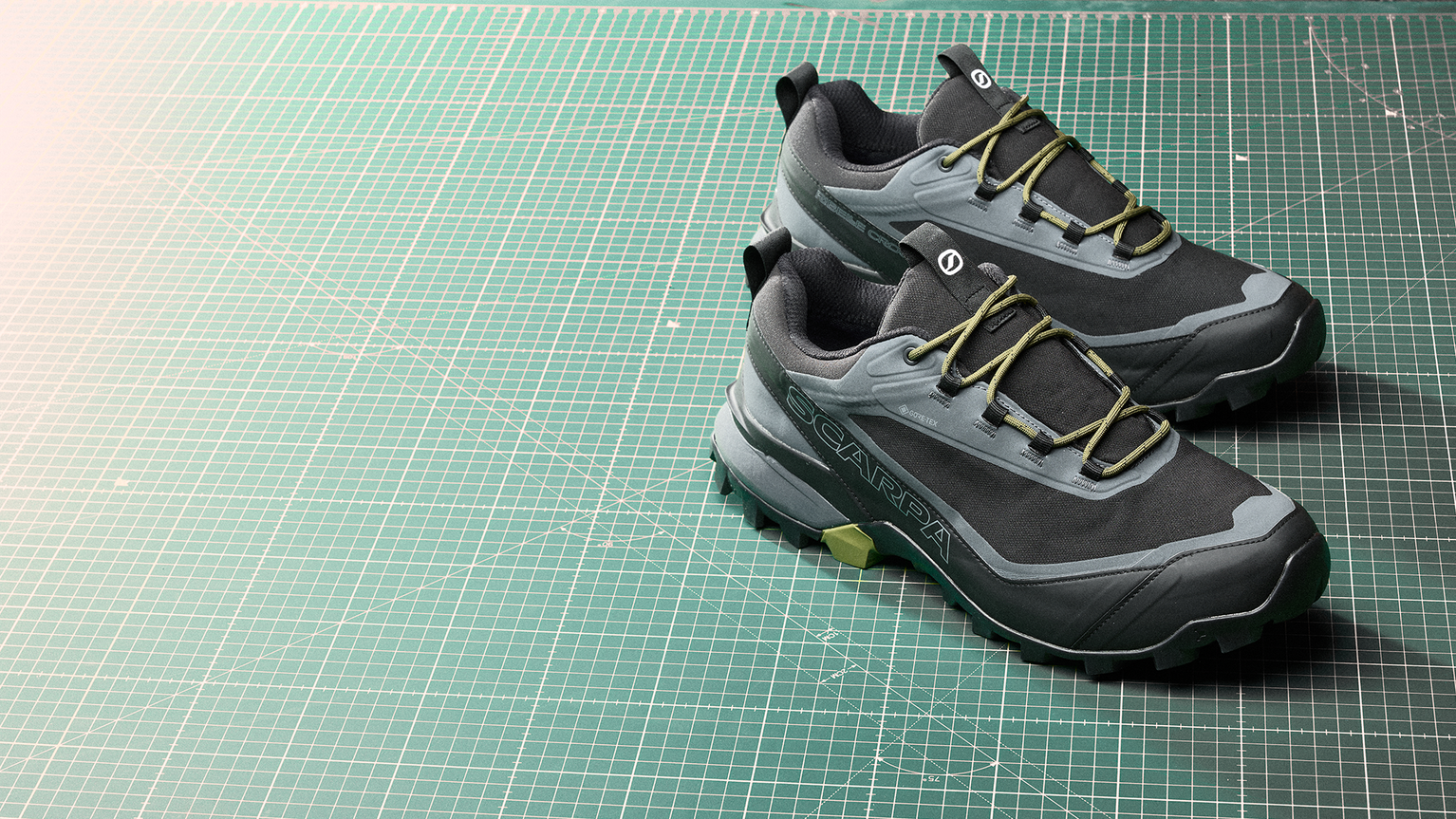 A pair of dark gray and green hiking shoes with yellow laces on a green cutting mat with grid lines.