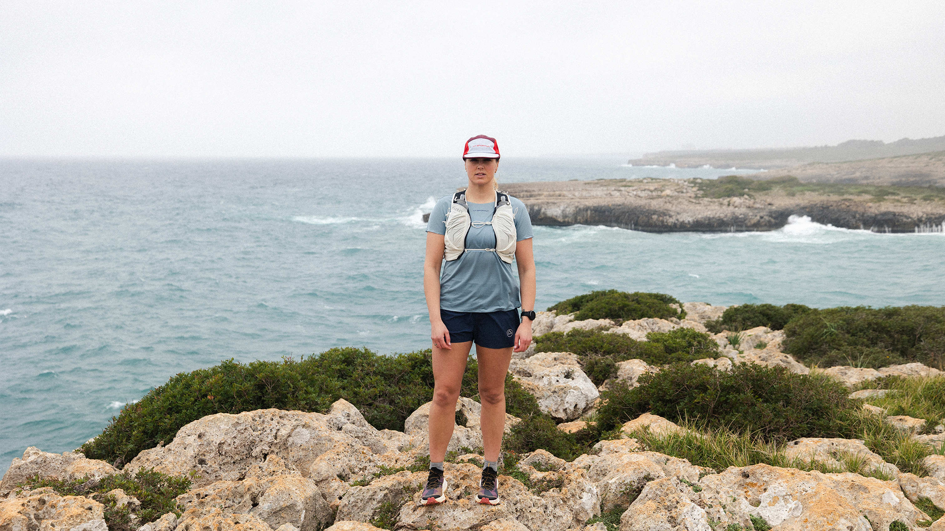 Person standing on rocky seaside cliff, wearing athletic gear and backpack, with ocean waves and cloudy sky in the background.