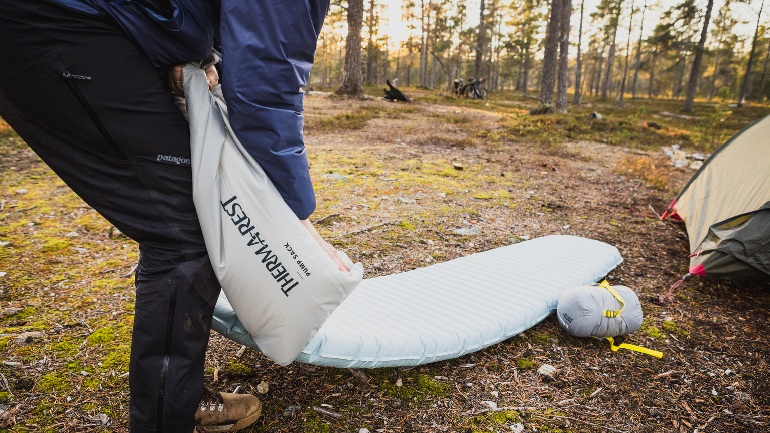 Person inflating a Therm-a-Rest sleeping pad in a forest campsite, with a tent and sunlight filtering through trees in the background.