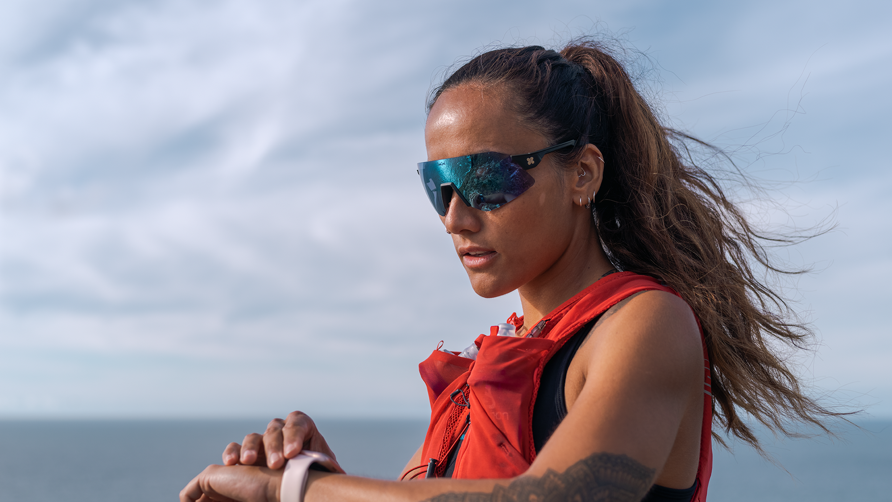 A woman in a red vest and sunglasses checks her smartwatch against a cloudy sky and ocean backdrop.