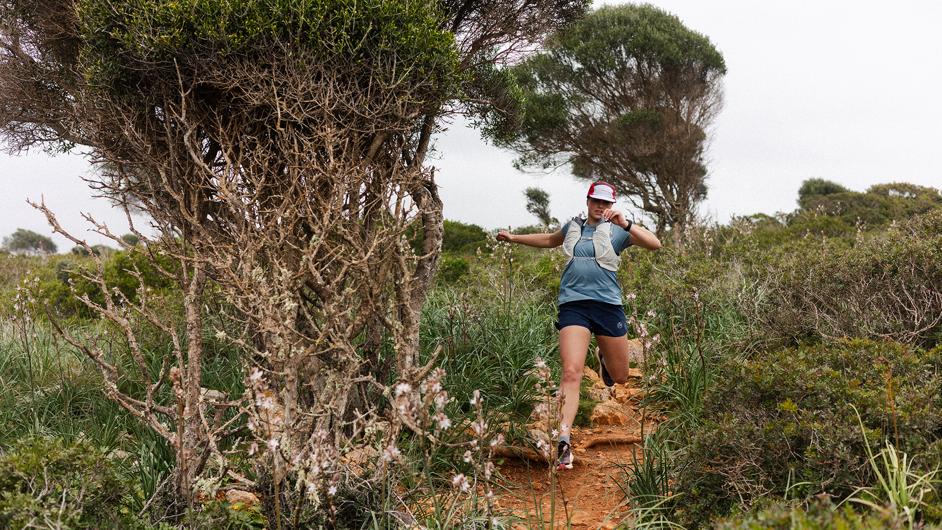 A person runs energetically down a rugged trail, surrounded by lush shrubs and trees, showcasing a dynamic mountain sports activity.