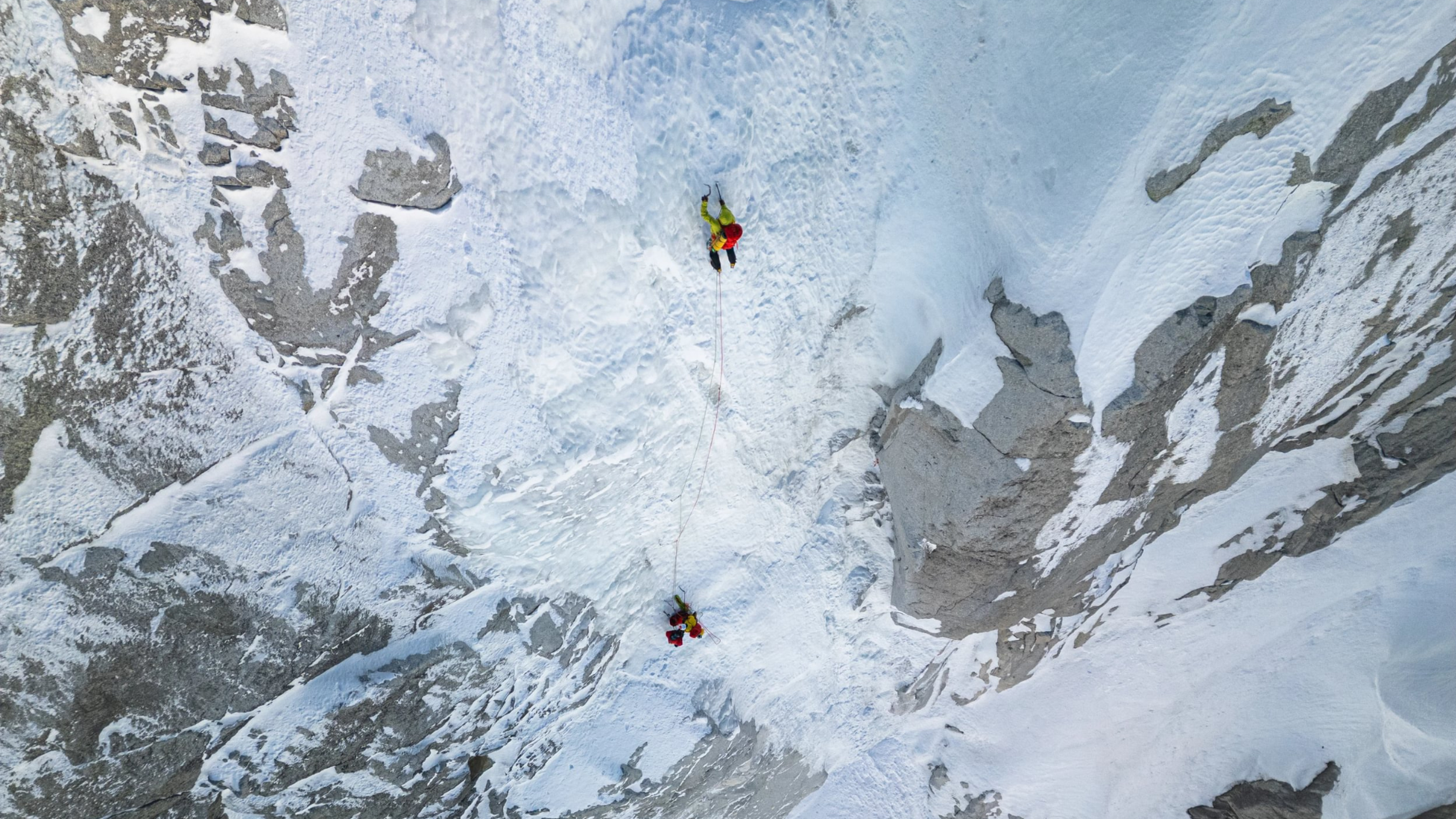 Two climbers in mountain equipment gear ascend a steep, snowy mountain face, connected by a rope, surrounded by rugged, icy terrain.