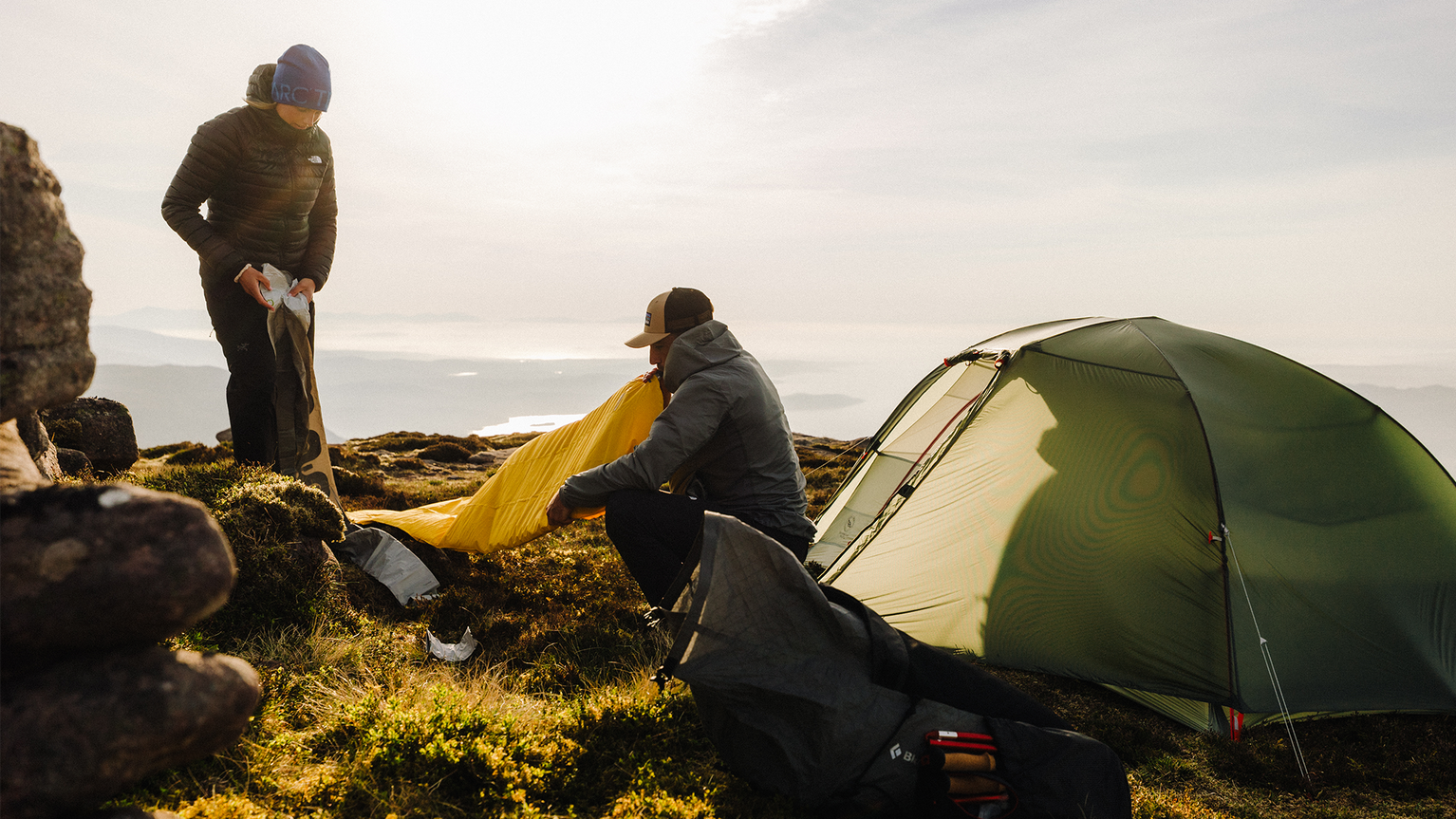 Two people setting up camp on a sunlit mountain, with a green tent and camping gear on grassy terrain.