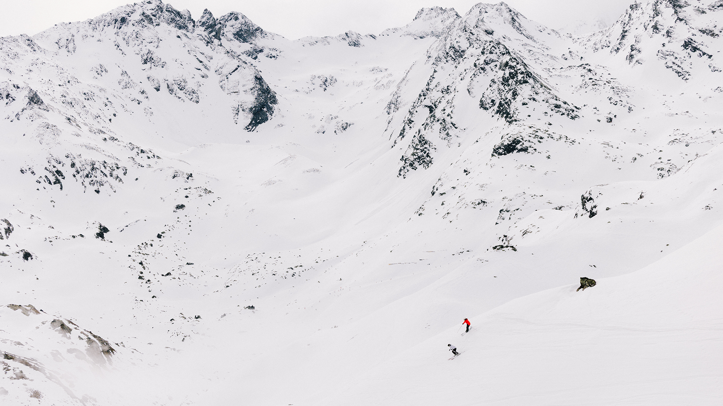 A skier in a red jacket descends a snowy mountain slope, surrounded by rugged, snow-covered peaks under a cloudy sky.