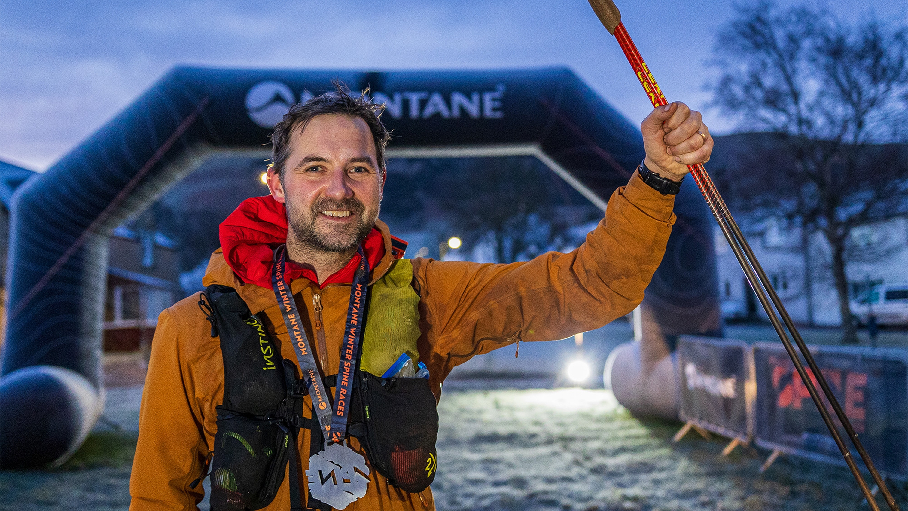 Person in an orange jacket holding walking poles, standing under a Montane archway at dawn, smiling confidently.