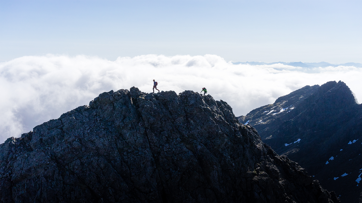Two climbers traverse a jagged mountain ridge under a bright sky, with vast clouds below and distant peaks in the background.