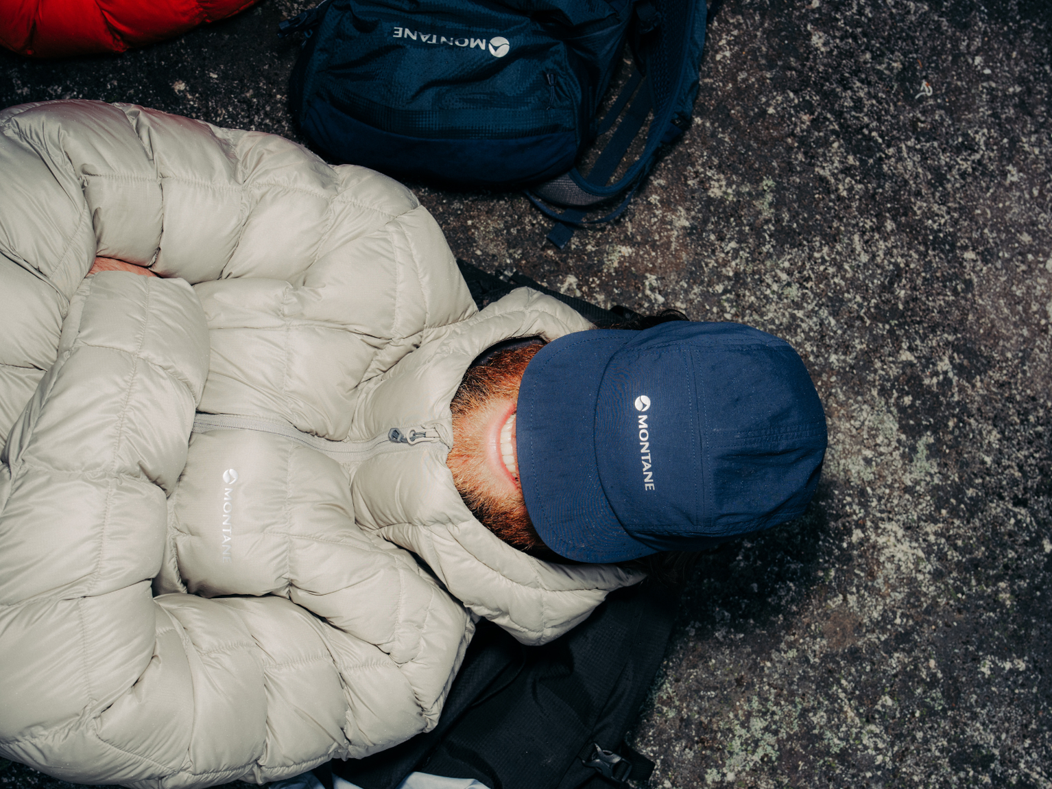 Person wearing a grey puffer jacket and a blue cap, lying on the ground next to backpacks, viewed from above.