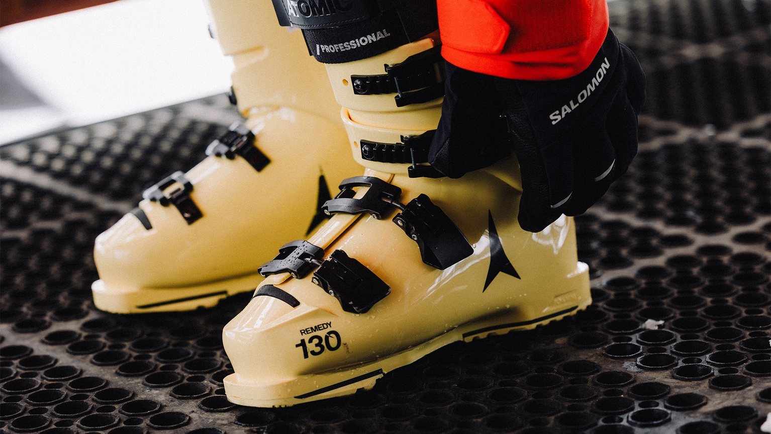 Close-up of yellow ski boots with black buckles on a textured mat. A gloved hand rests on one boot, featuring the brand name "Salomon."