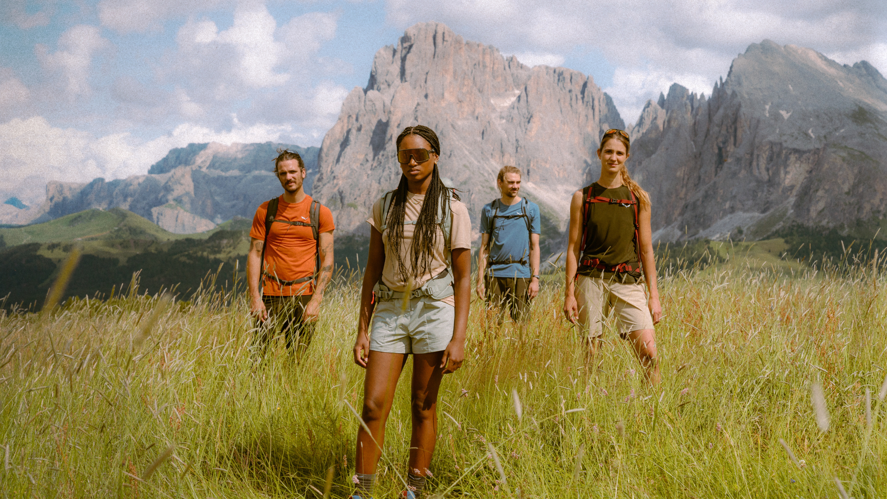 Four people hiking in a grassy field with backpacks, mountains in the background, under a partly cloudy sky.