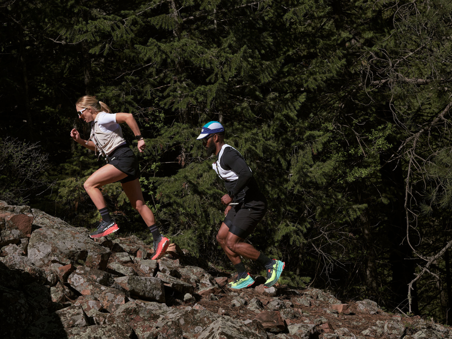 Two people trail running uphill on rocky terrain, surrounded by dense green forest. Both wear athletic gear and appear focused.