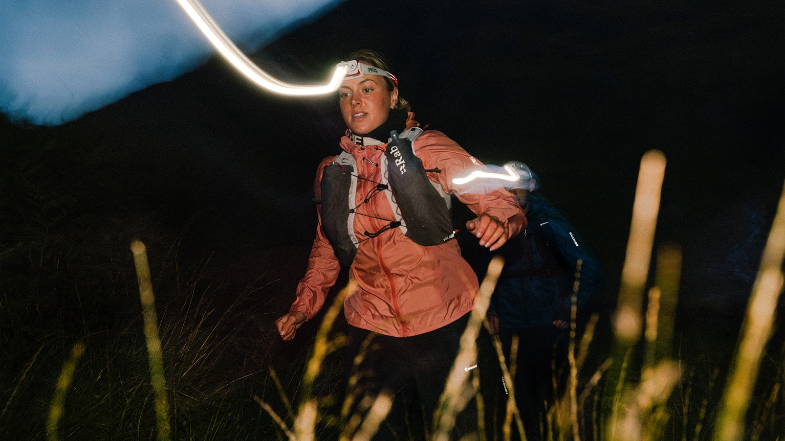 A woman trails through a dimly lit path, wearing a headlamp and mountain gear.