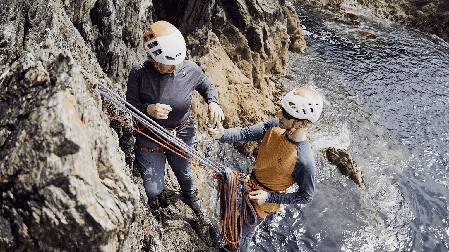 Two climbers wearing helmets and climbing gear, exchanging a carabiner on a rocky cliff above the sea.