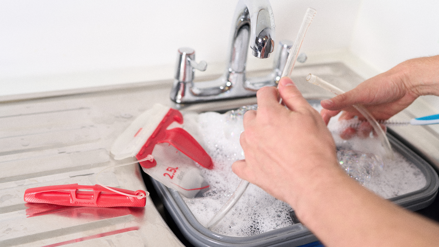 Person washing a baby bottle and accessories in a sink filled with soapy water, with a red bottle brush and drying rack nearby.